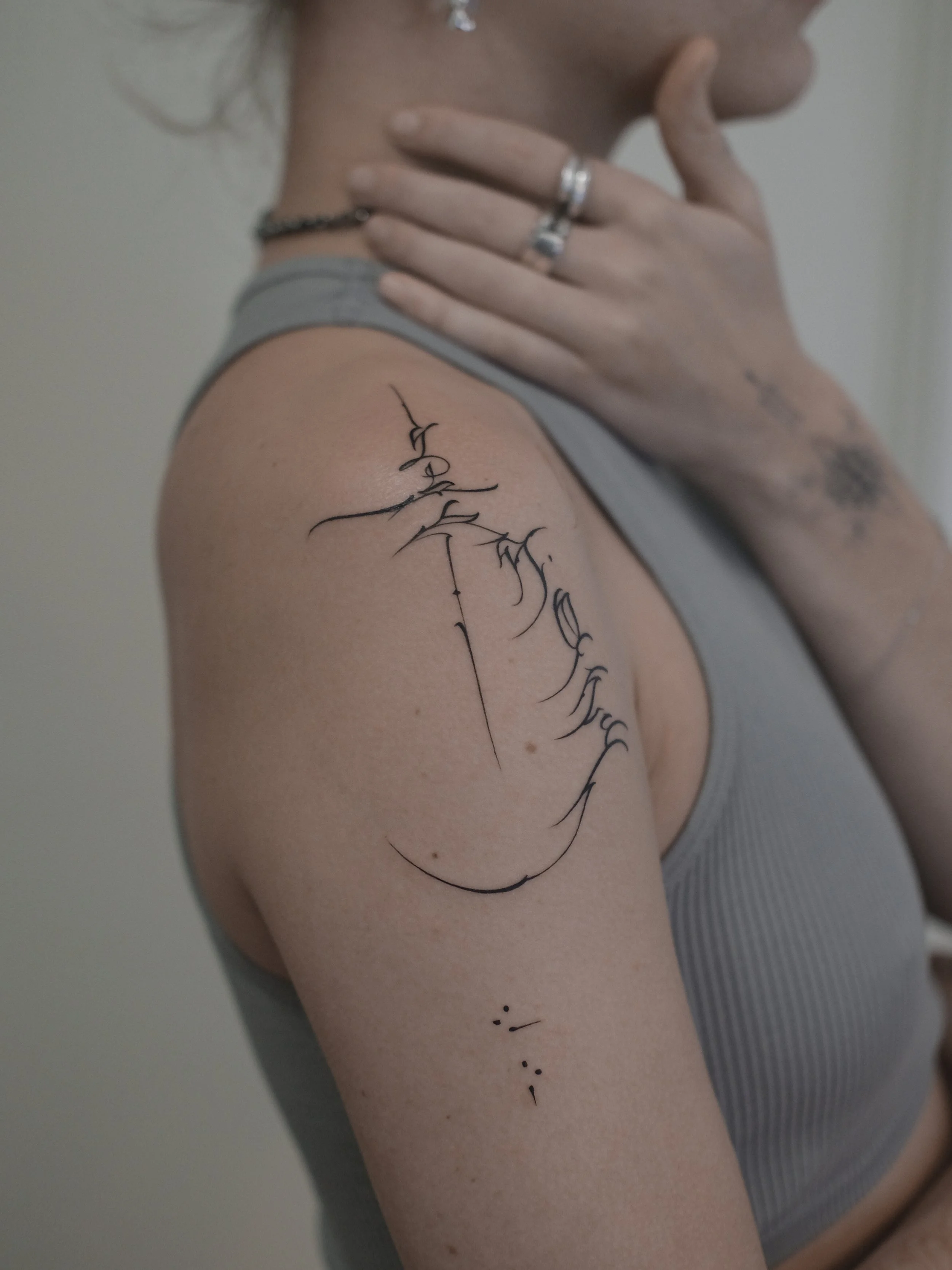 Close-up of a person's shoulder and upper arm with a black tattoo of flowing cursive text. The person is wearing a gray sleeveless top, silver rings, a bracelet, and a necklace. The background is plain and light-colored.