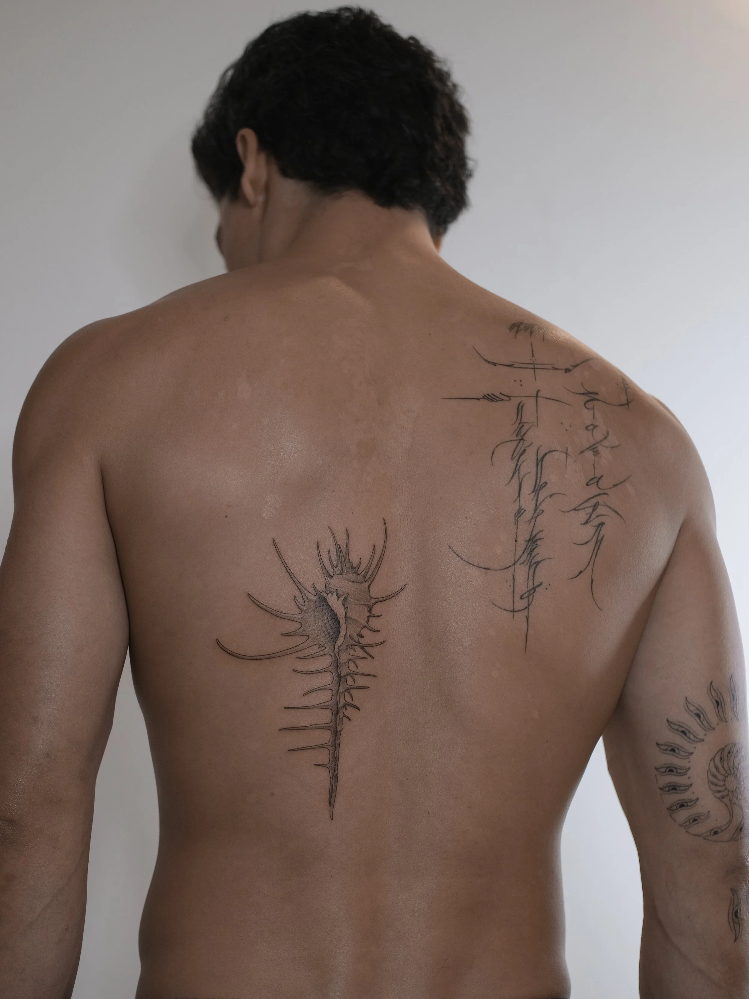 Back view of a person with short dark hair, showing a tattoo of a spine and abstract lines on their upper back and shoulder.