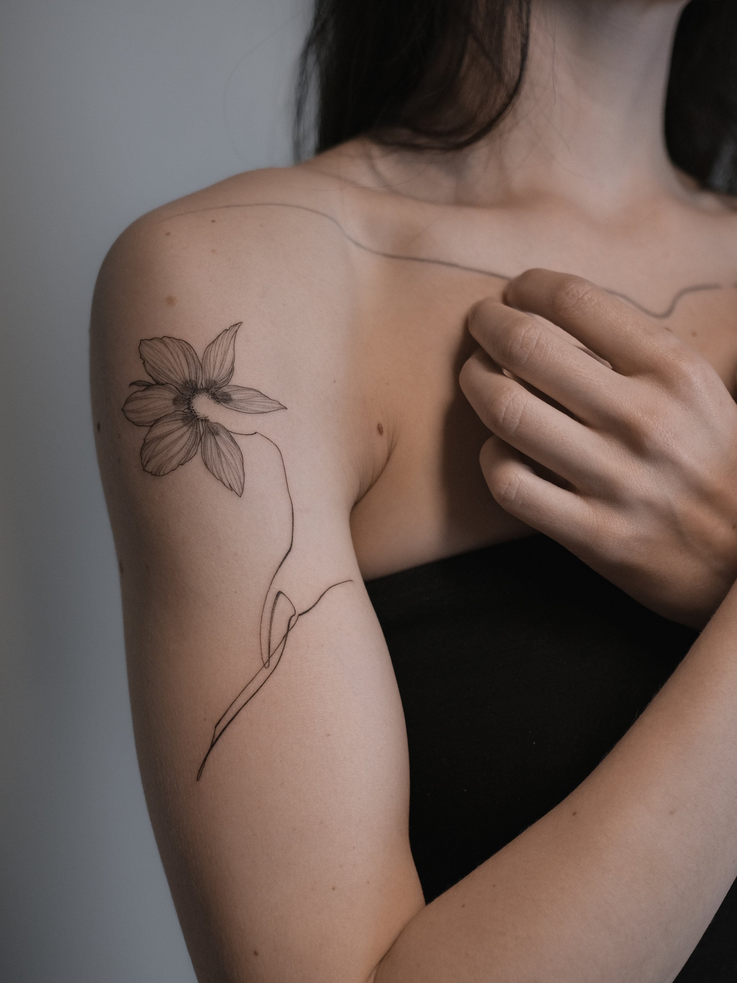 A close-up of a woman's shoulder and upper arm, featuring a tattoo of a flower with five petals and a long, flowing stem. The woman has fair skin and dark hair.