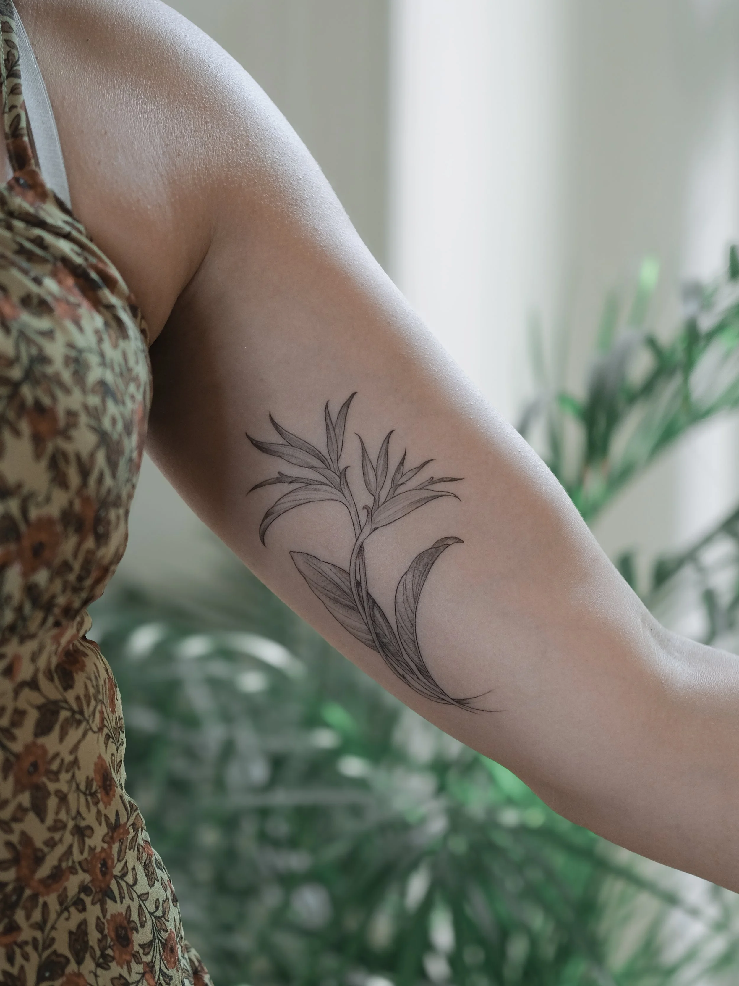 Close-up of a person's arm with a floral tattoo, wearing a floral patterned dress, with green plants in the background.