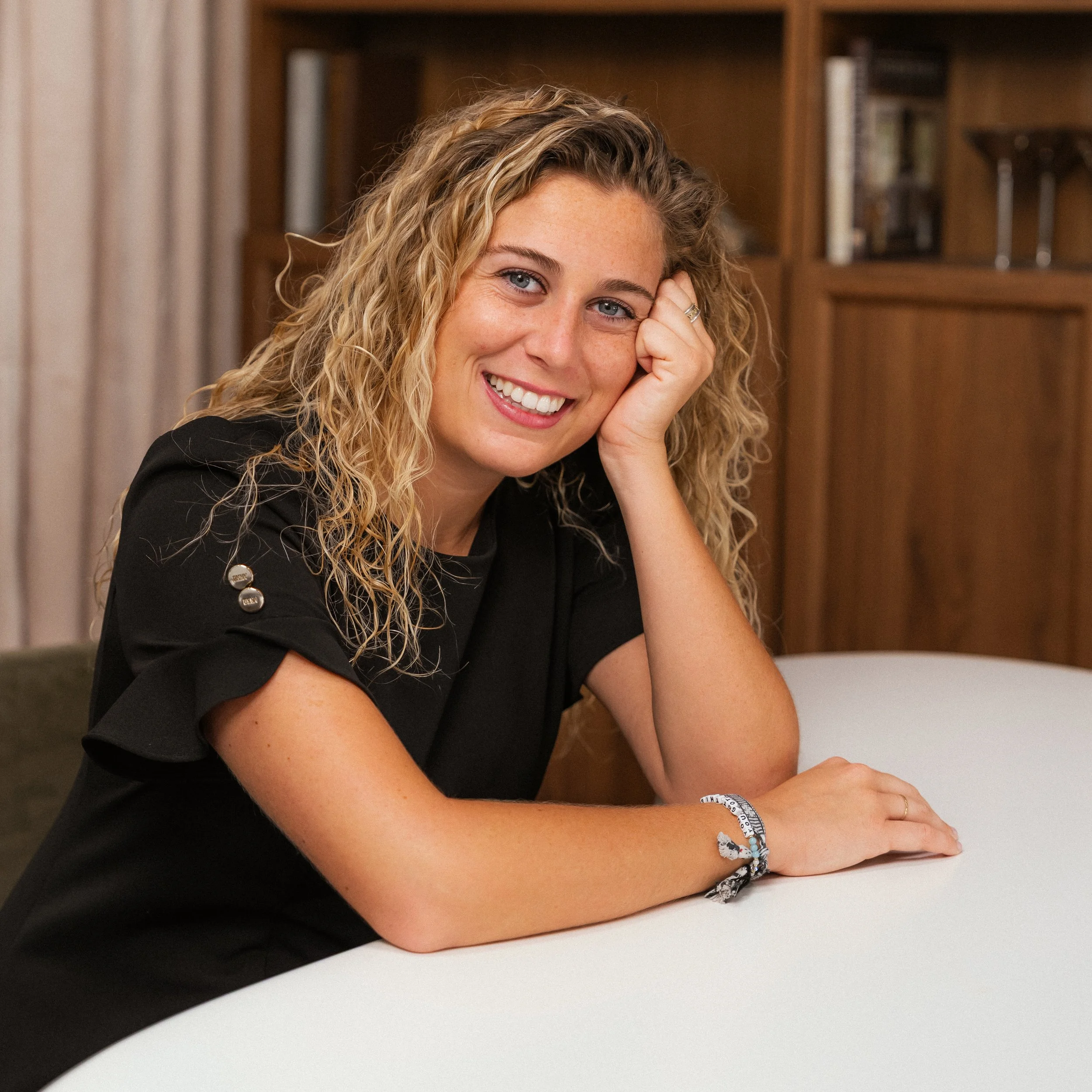 A smiling woman with curly blonde hair resting her head on her hand at a table, wearing a black top and jewelry, in a room with wooden furniture.