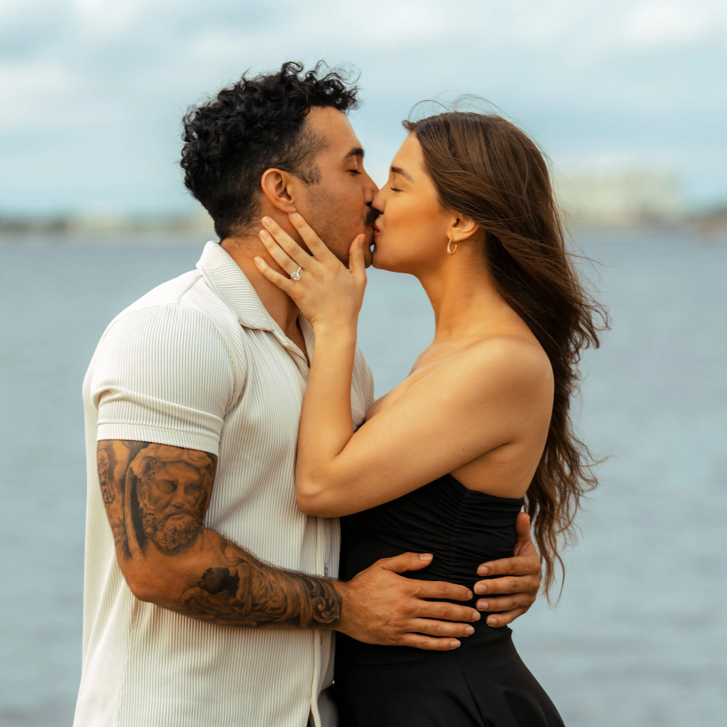 A couple kissing by the water outdoors, with the man wearing a white short-sleeved shirt and the woman in a black strapless dress.