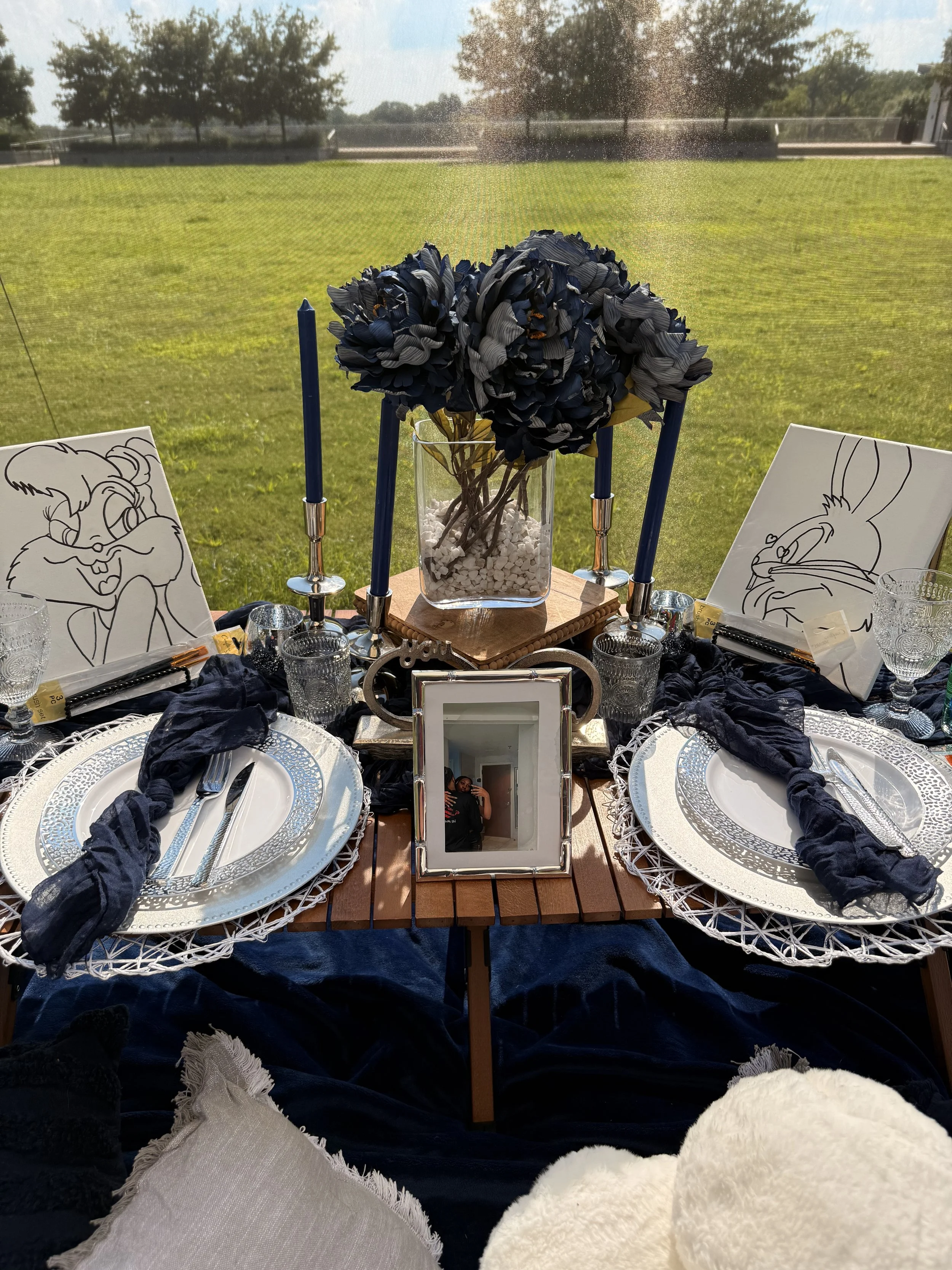A decorated outdoor dining table with a flower centerpiece, framed photograph, and cartoon sketch artwork, set with plates, glasses, napkins, and cutlery.