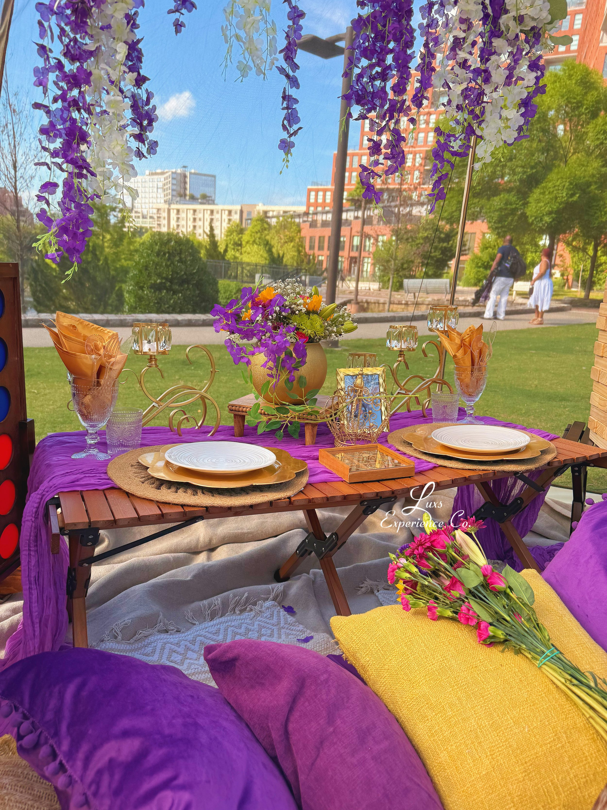 A colorful outdoor picnic table decorated with purple and yellow pillows, a bouquet of pink flowers, and a purple table runner, with a city park background featuring tall buildings and green trees.
