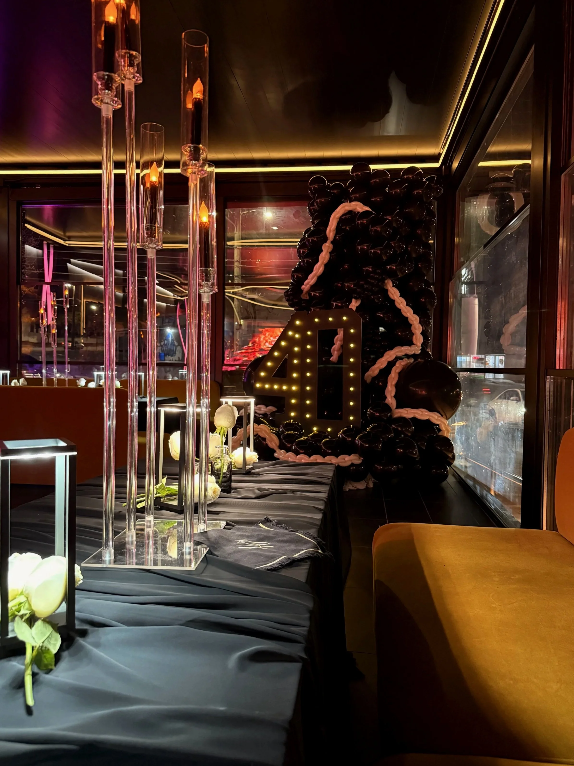 Decorated table with white roses, candleholders, and a black tablecloth, near a large balloon arrangement with a large illuminated number '40' sign, in a dimly lit room with dark walls and large windows.