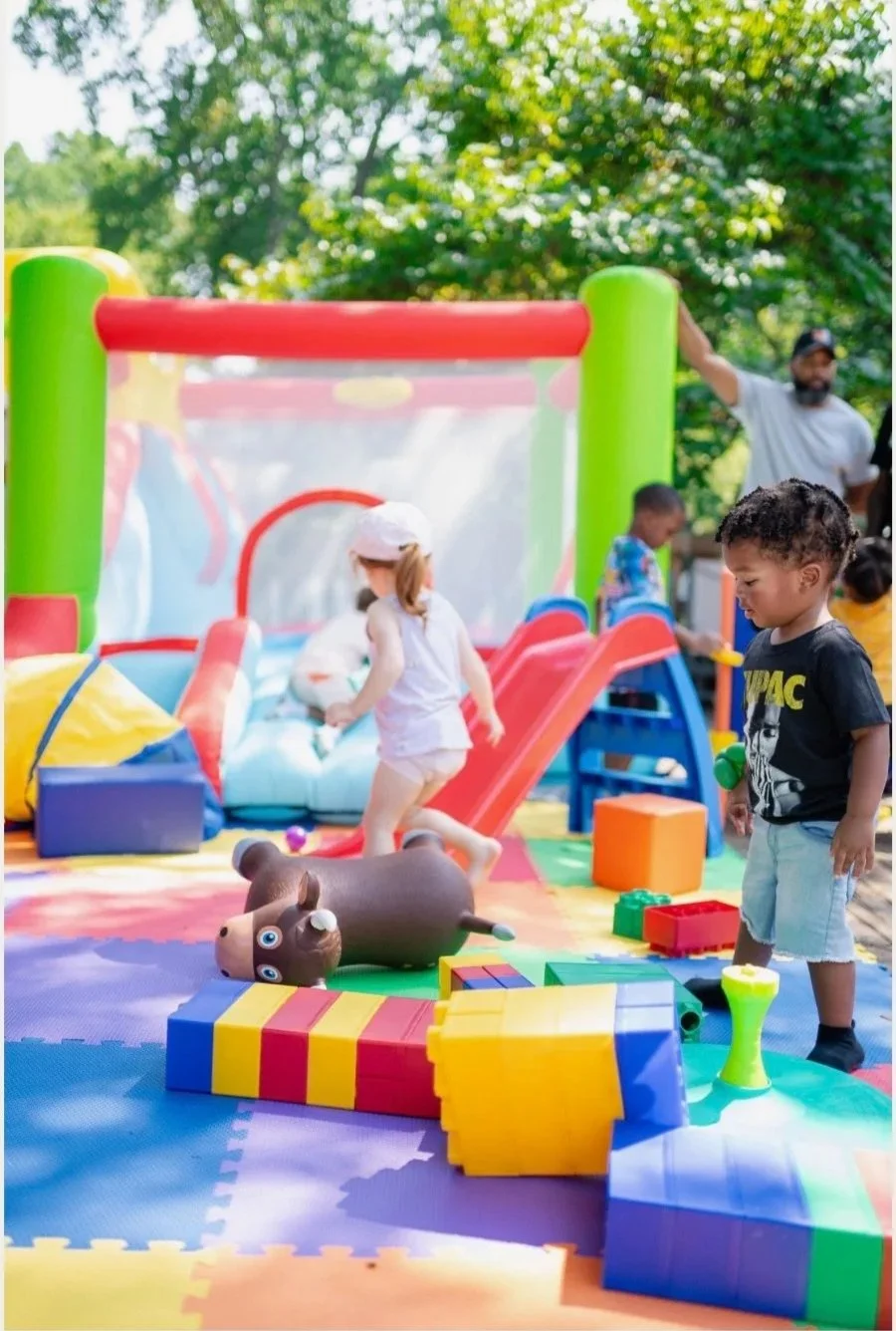 Children playing in an outdoor bounce house and toy area with colorful foam blocks and trees in the background.