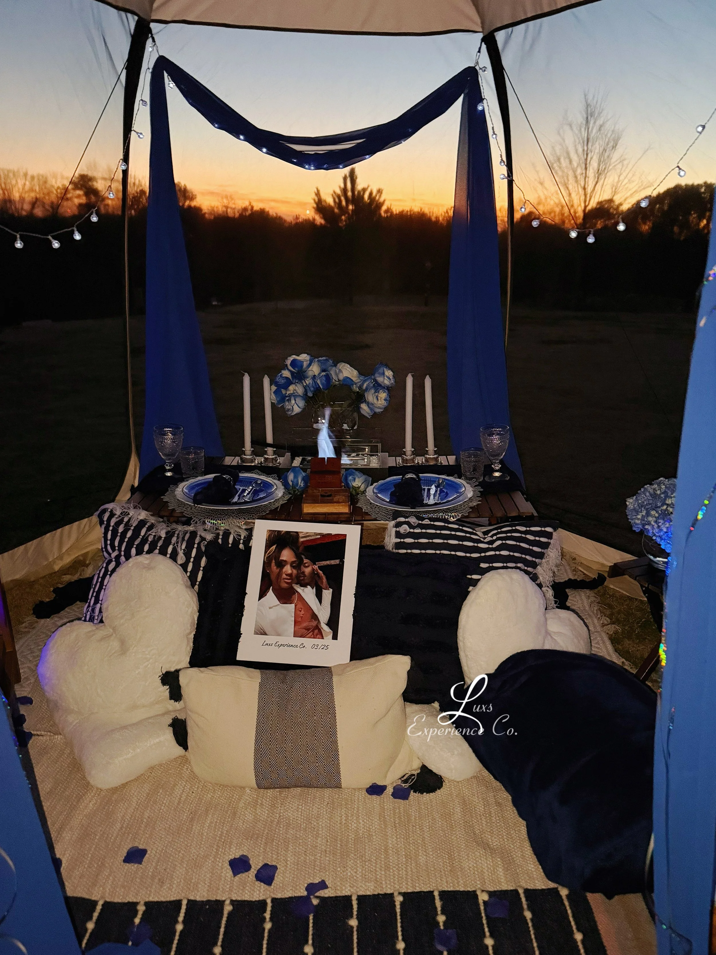 A cozy outdoor dining setup inside a tent at sunset, featuring a table with blue and white plates, candles, and a floral centerpiece, surrounded by plush pillows and teddy bears, with ambient string lights and a photograph of a woman on a black cloth
