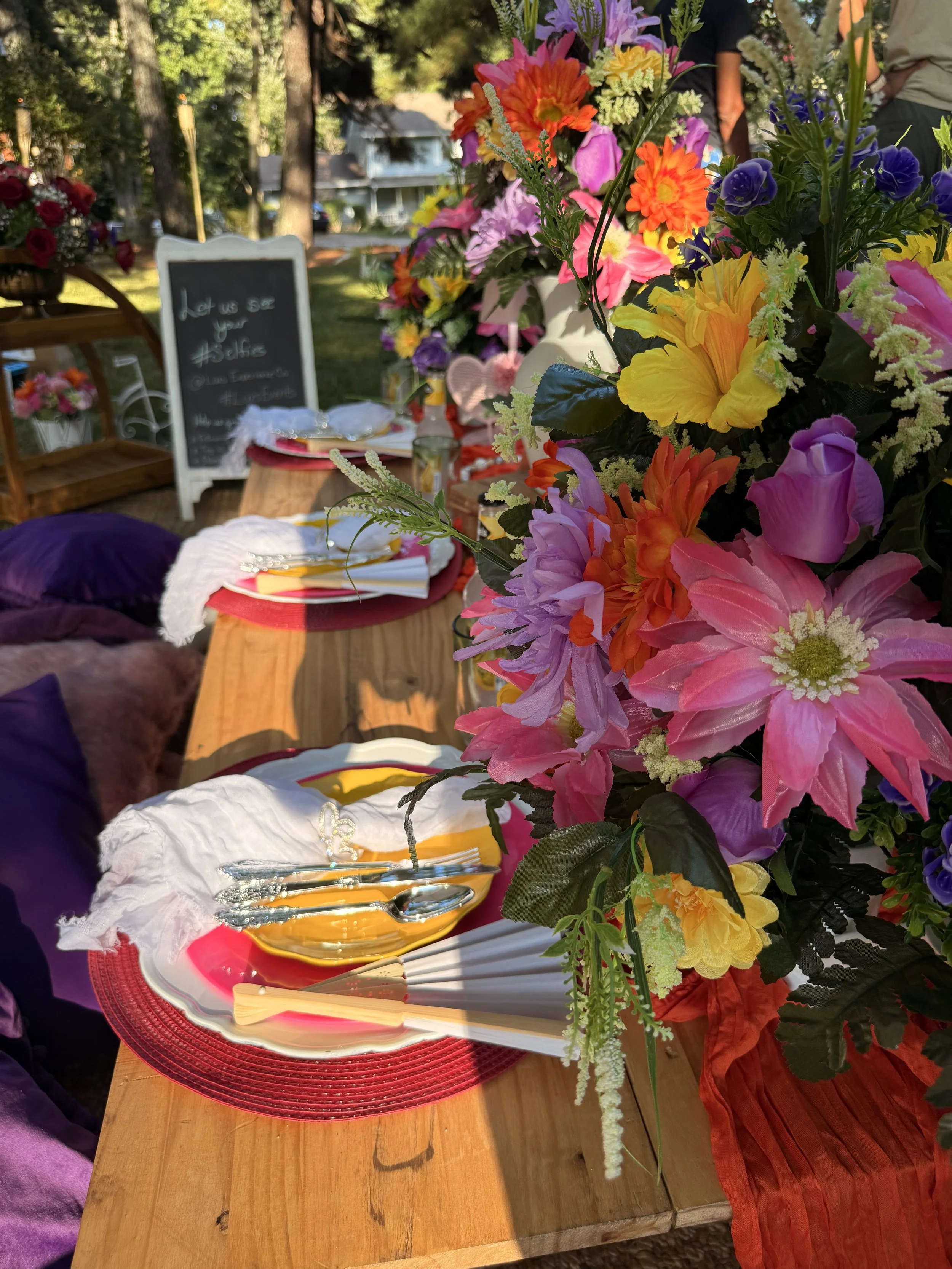 A decorated outdoor table with colorful floral centerpiece, plates, utensils, and napkins for a gathering or celebration.