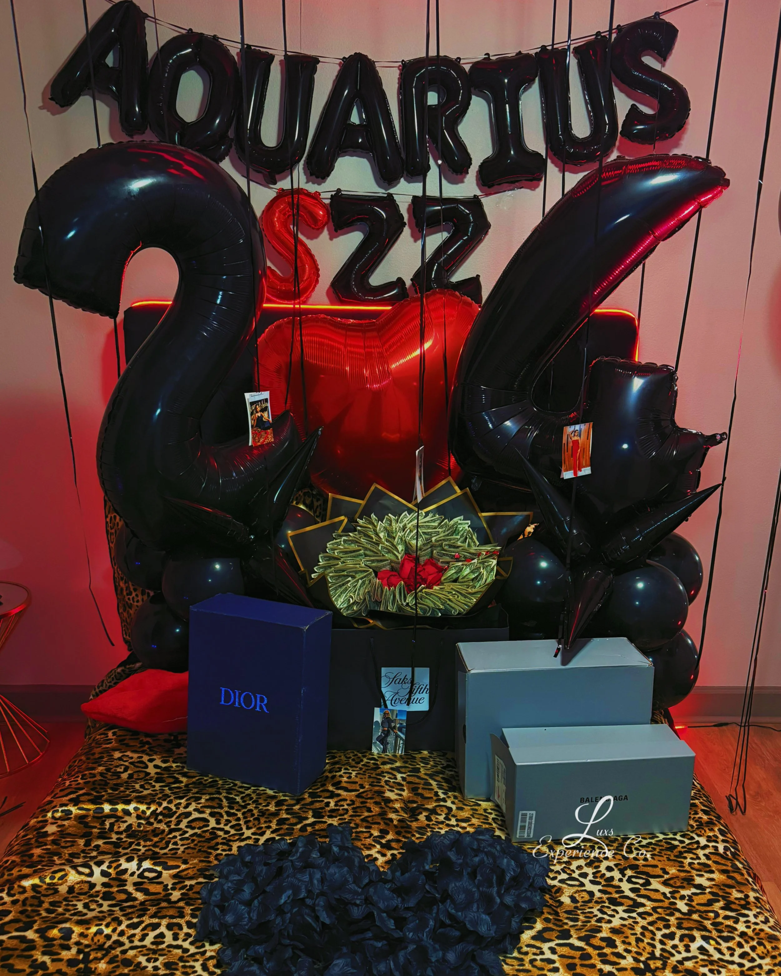 Black and red balloons spelling 'YouAre1S2', with large black balloons displaying '2022'. Gift boxes from Dior and Balenciaga on a leopard print cloth, with a black floral decoration and a framed photo, amid red lighting.
