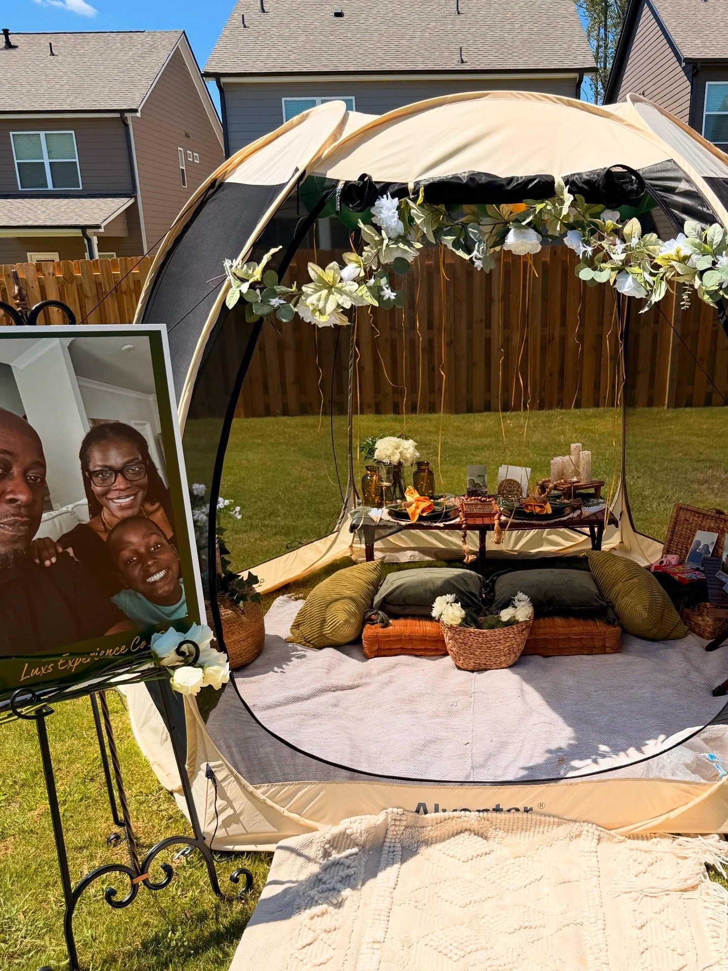 A backyard setup with a beige outdoor shelter decorated with a floral garland, featuring a table with candles, flowers, and decorative items, surrounded by cushions and baskets. A large wedding photo of an African American family is displayed on a st