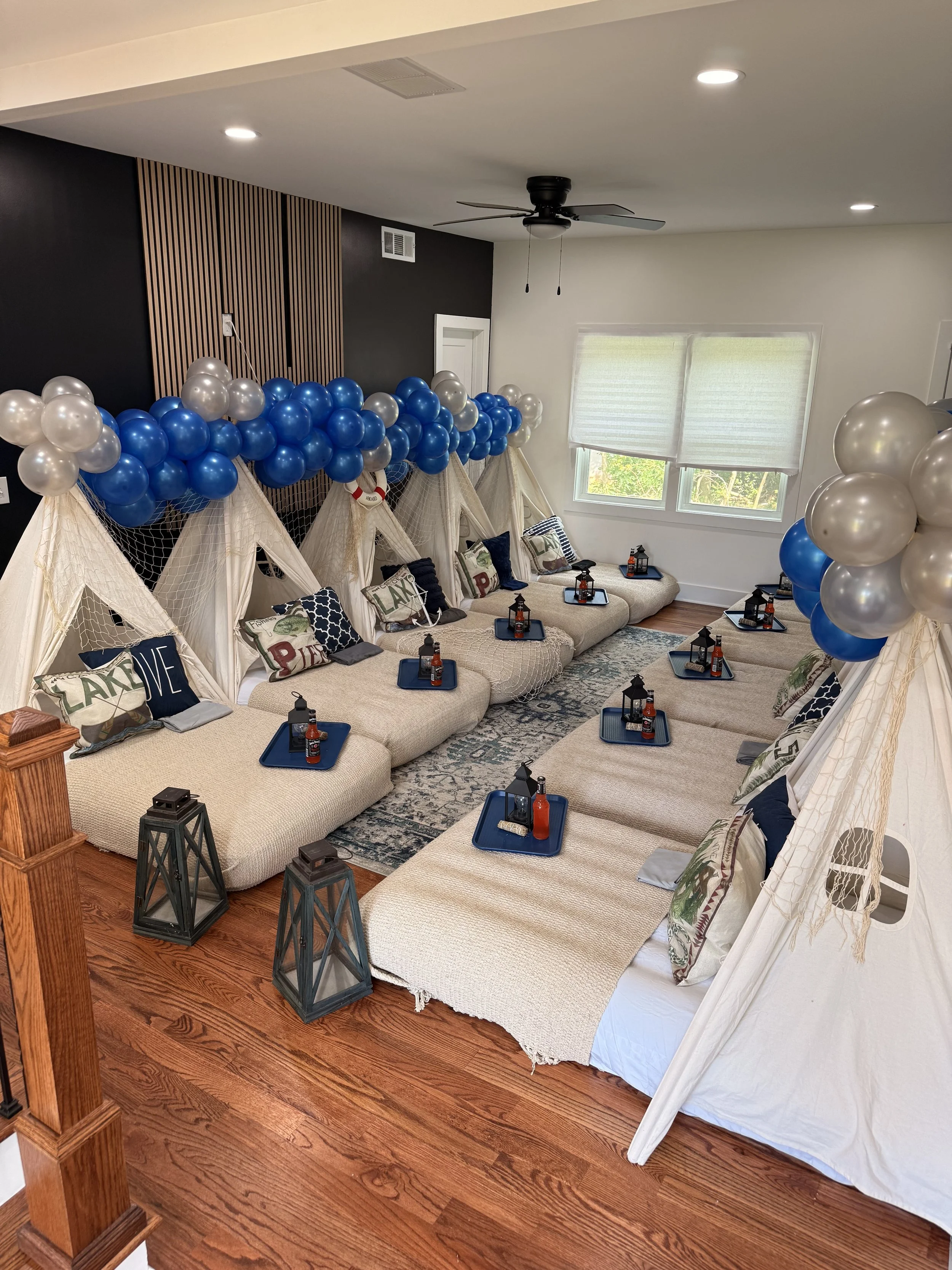 Indoor setup with small beige sofas arranged in a U-shape, decorated with navy and beige pillows, each with a tray holding a bottle of ketchup, napkin, and condiment bottle. The room features white walls, a window with blinds, a black ceiling fan, an