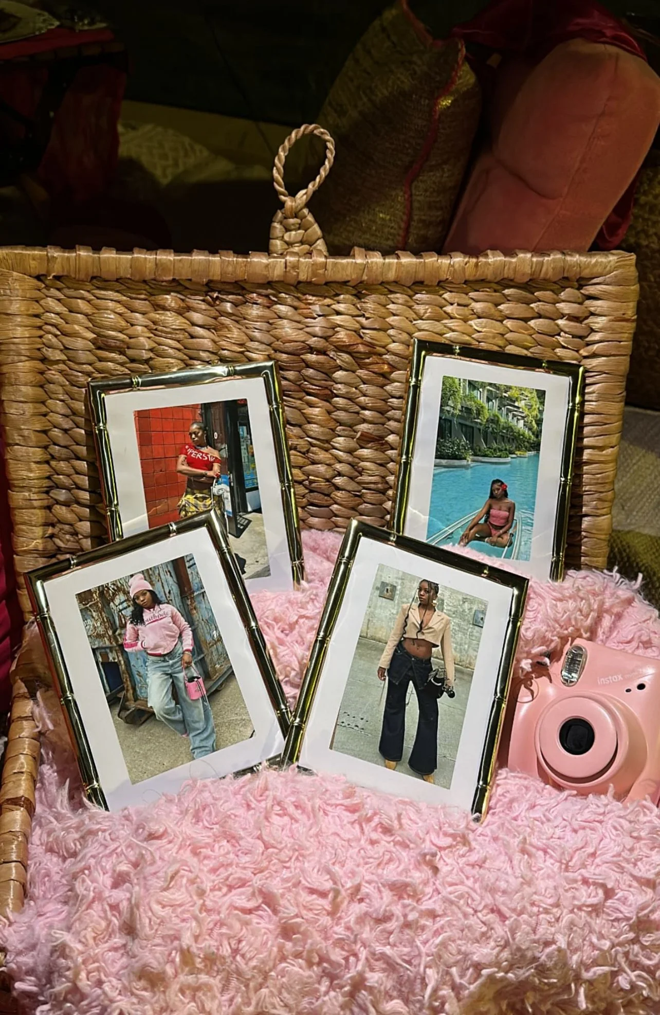 A wicker basket holding five framed photographs of a woman in various outfits and settings, with a pink Instax camera nearby, placed on pink fluffy fabric.