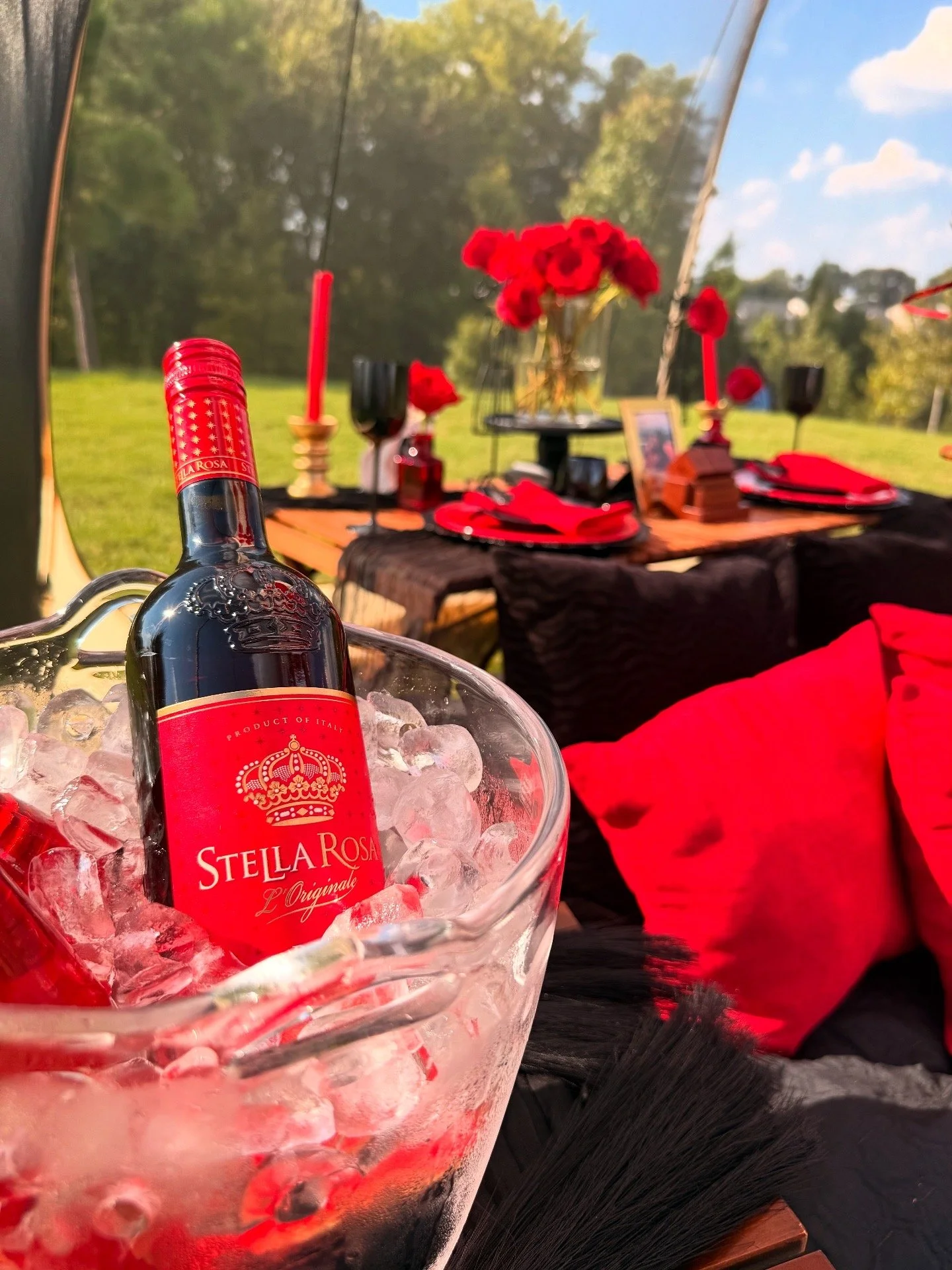 Elegant outdoor table set with white ornate plates, gold utensils, beige napkins with decorative bows, gold and clear glasses, and a rustic burlap table runner, decorated with white flowers and candles for a celebration or special event.