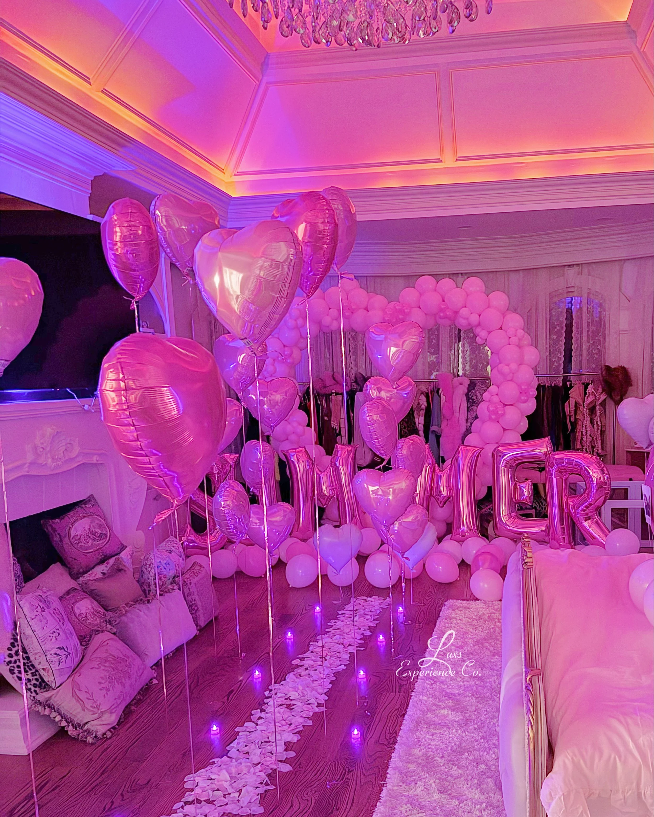 A room decorated with pink heart-shaped balloons, a balloon arch, and large pink letter balloons spelling "MOM". Pink and white balloons cover the floor and walls, with a heart-shaped balloon garland in the background. Soft pink lighting enhances the