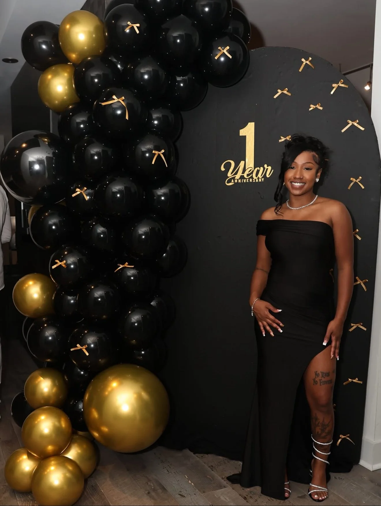 A woman in a black sleeveless dress standing next to a black and gold balloon arch with a black backdrop that reads '1 Year Anniversary.' The woman is smiling, wearing jewelry, and showing a tattoo on her thigh. There are gold and black balloons deco