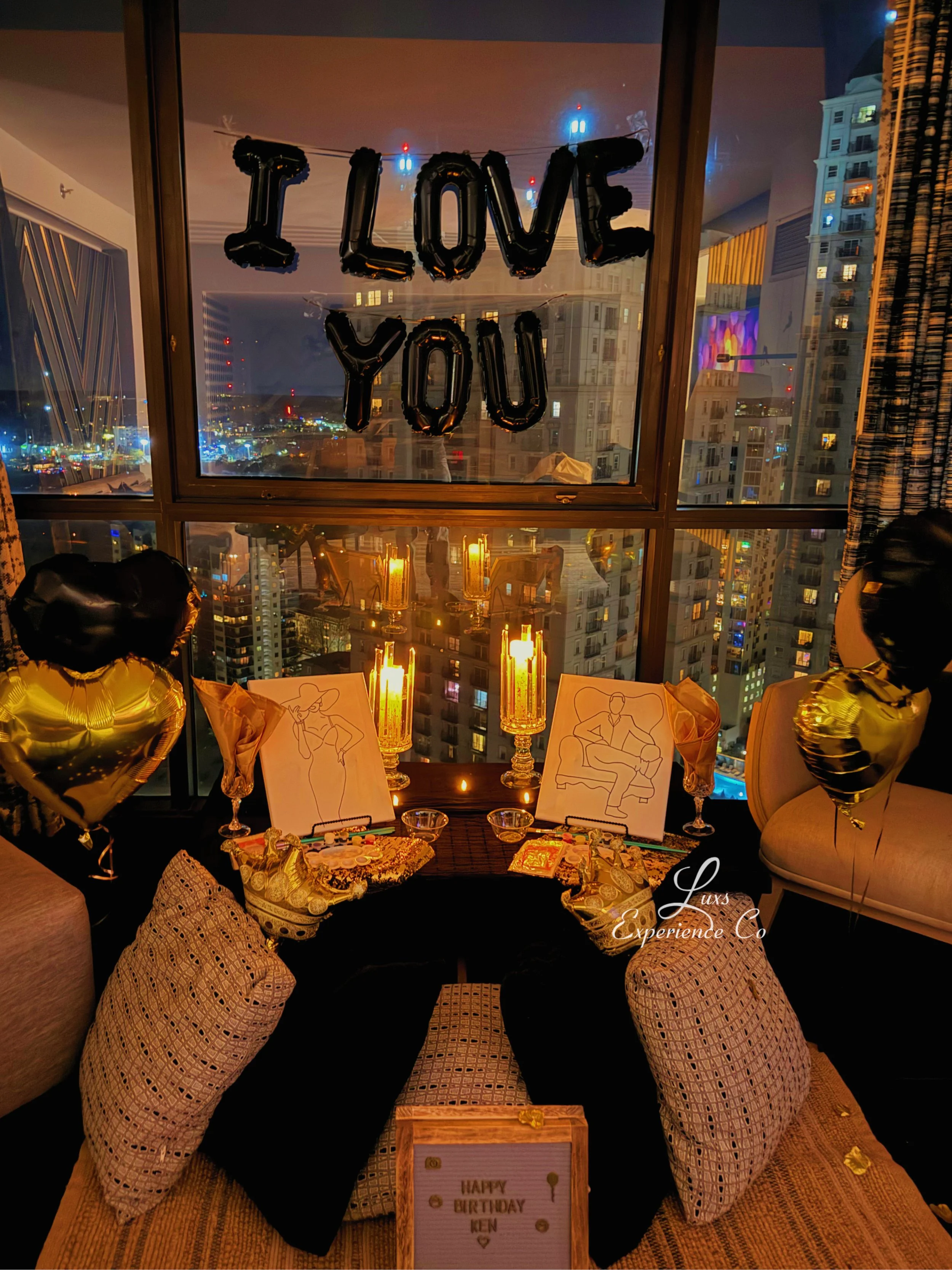 A birthday celebration setup in a city high-rise apartment with a nighttime cityscape view. There are black balloons spelling out 'I LOVE YOU' on the window, candles, artistic sketches, decorative pillows, heart-shaped balloons, and a small sign that