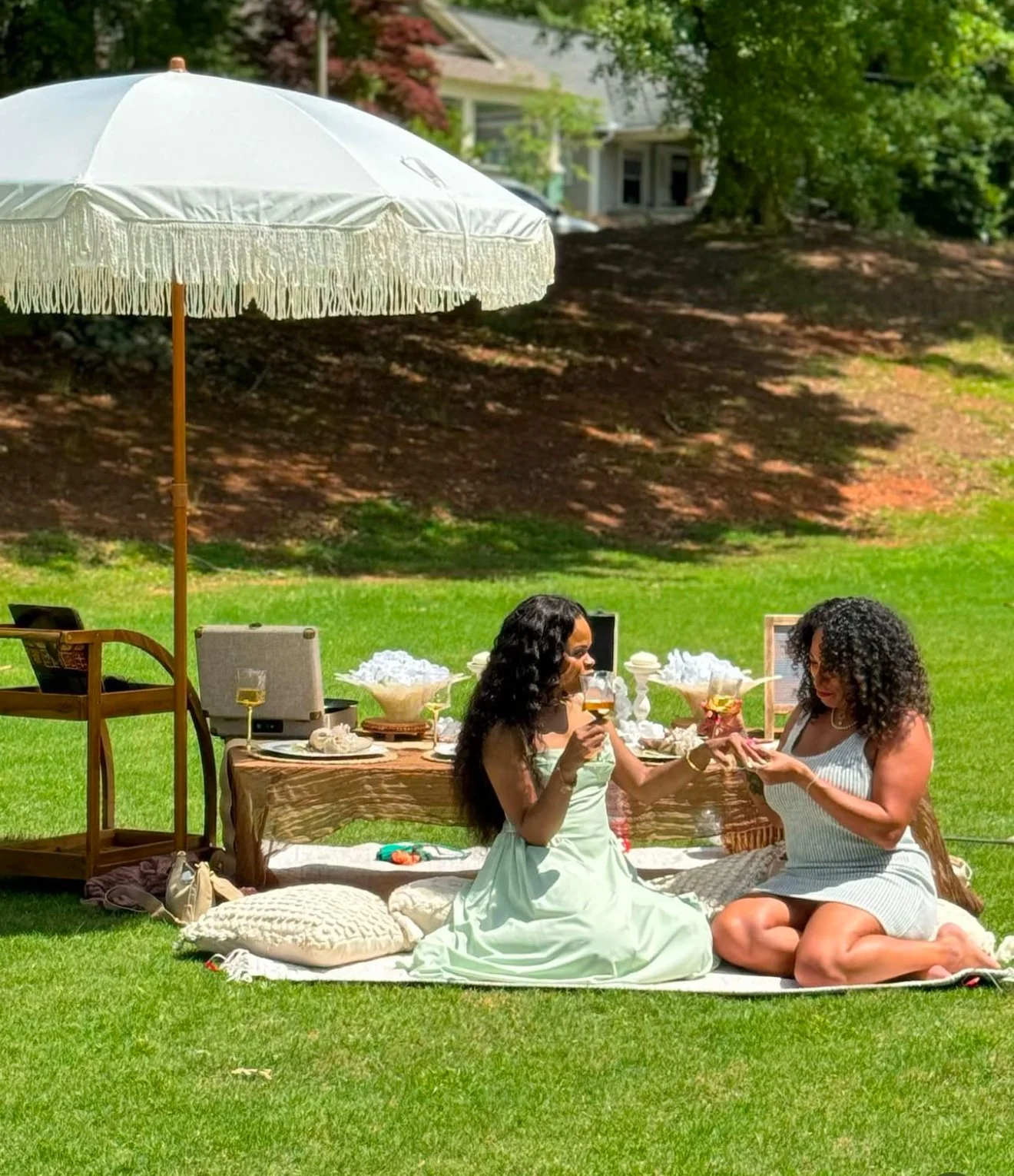 Two women having a picnic on a grassy lawn in Atlanta with a white umbrella, sitting on cushions in front of a low wooden table with snacks and drinks, engaging in a gift exchange or toast.