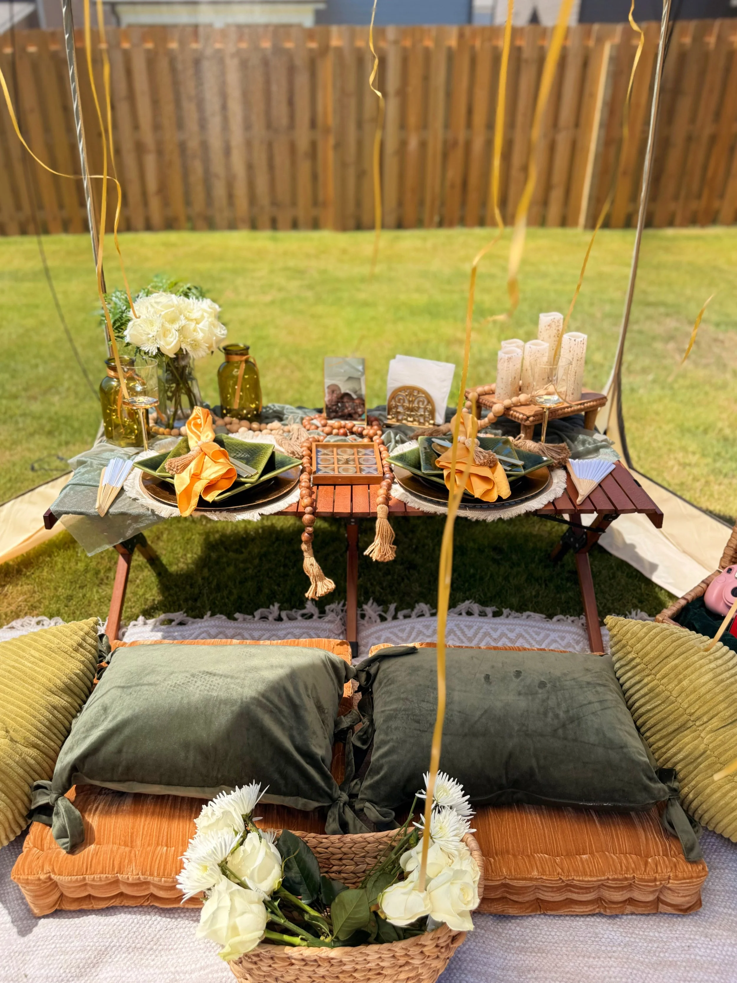 Outdoor decorated table with pillows, flowers, candles, and snacks arranged for a gathering or celebration in a backyard.