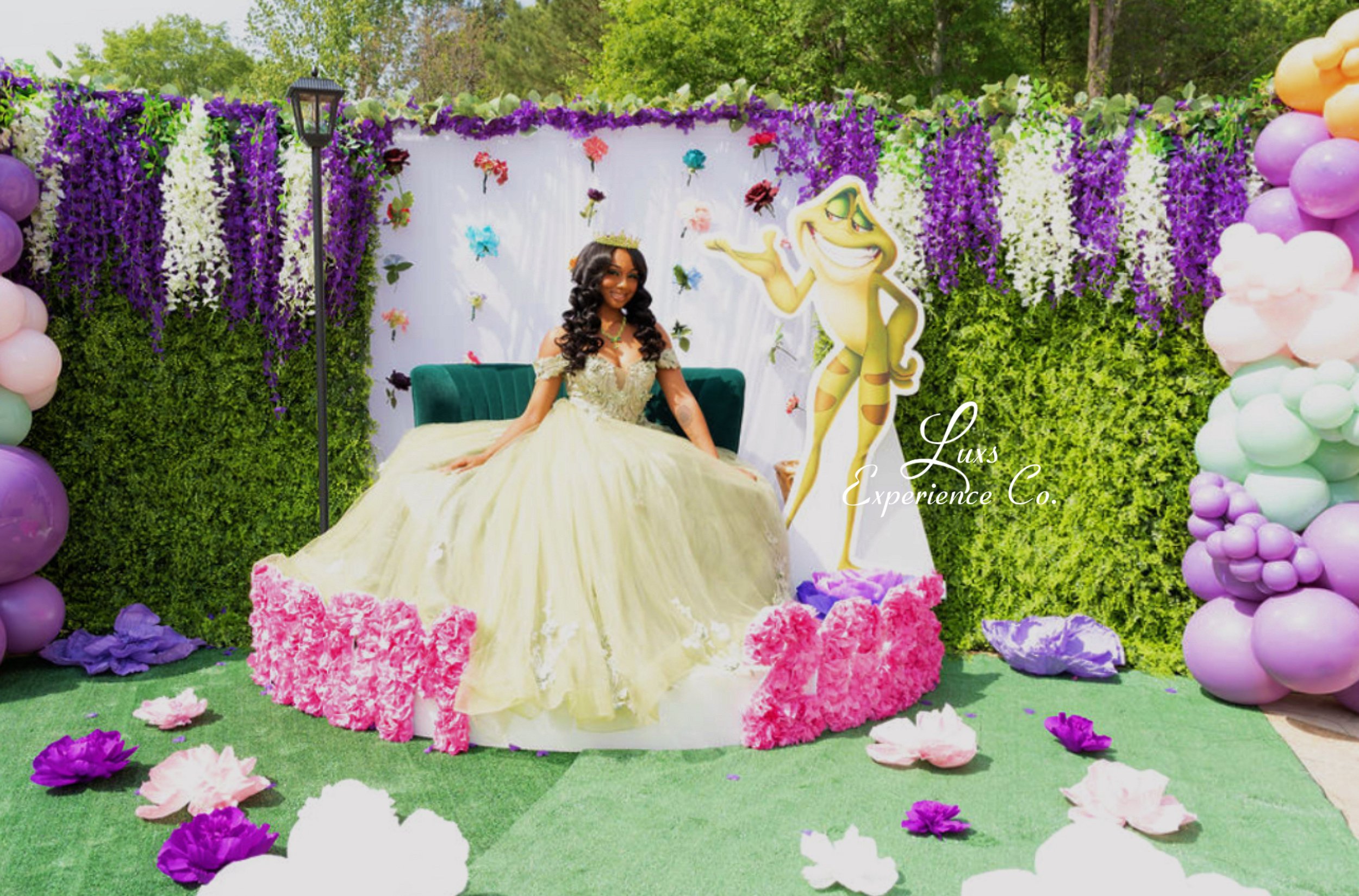 A young woman in a yellow ball gown seated on a green velvet sofa amidst a colorful birthday or celebration setup. The background features a white display wall decorated with purple and white flowers and butterfly accents. There is a large cartoon cu