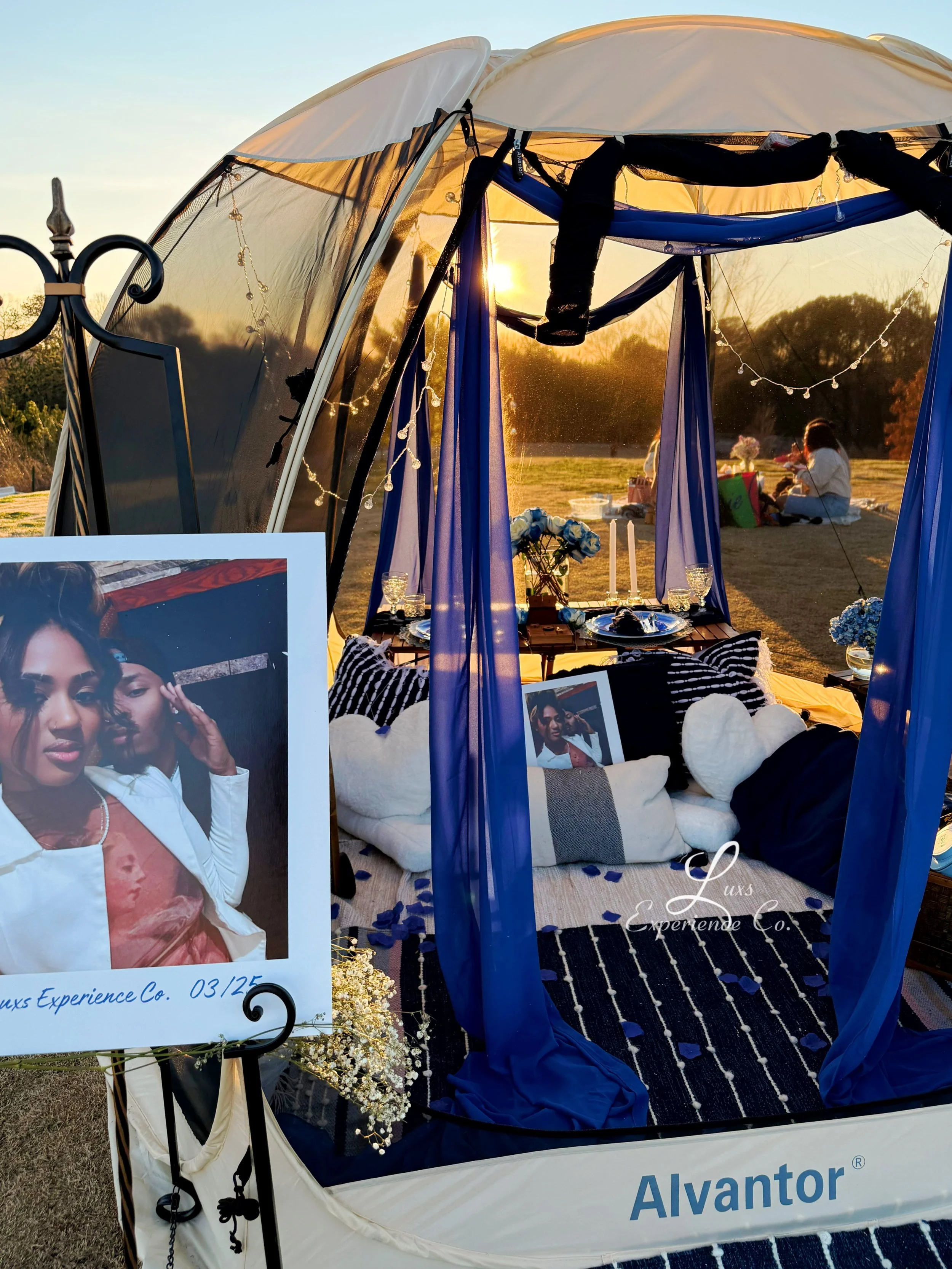A decorated outdoor tent with a table set for a romantic dinner, fairy lights, blue curtains, and pillows, with a poster of a woman in the foreground and people sitting outside in the background during sunset.
