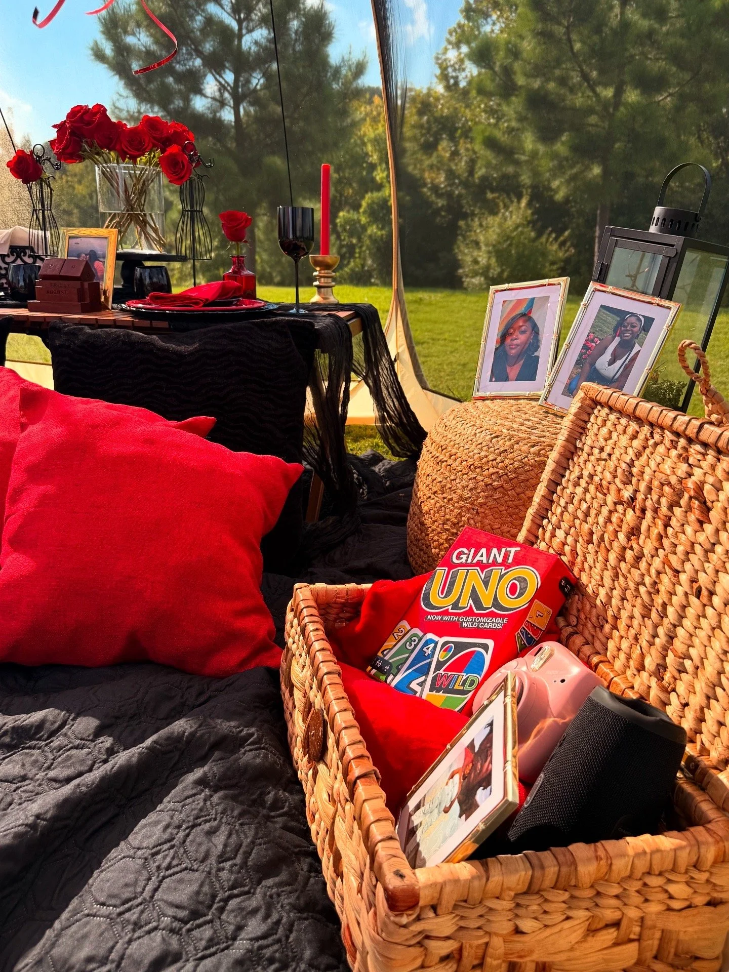 Outdoor picnic setup with red pillows, a basket of games, framed photos, a table with candles and flowers, and a lantern, all on a grassy field with trees in the background.