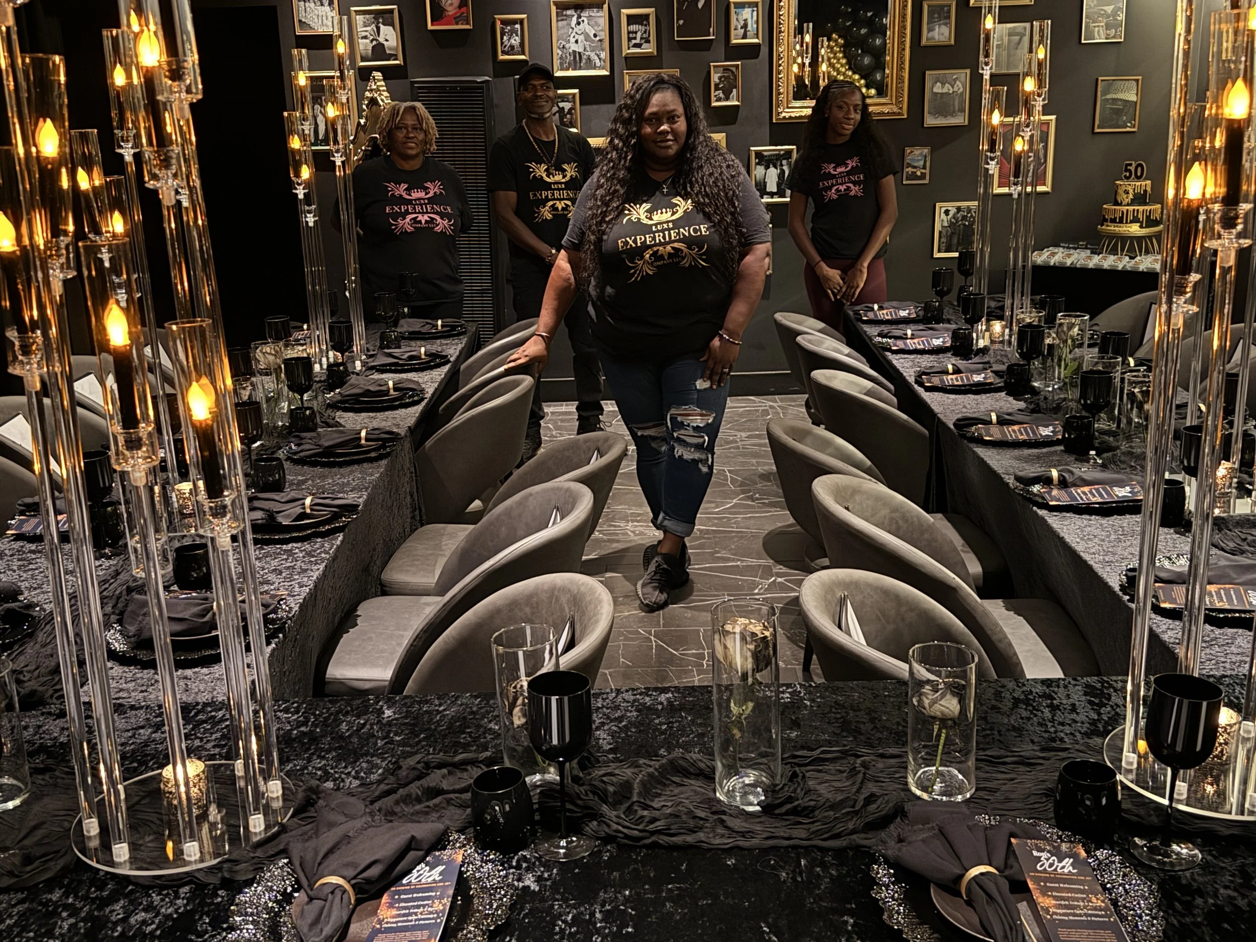 Group of five people wearing black shirts with gold lettering and designs, standing in a decorated dining room with black tablecloths, candles, and framed photos on dark walls, celebrating a 50th birthday.