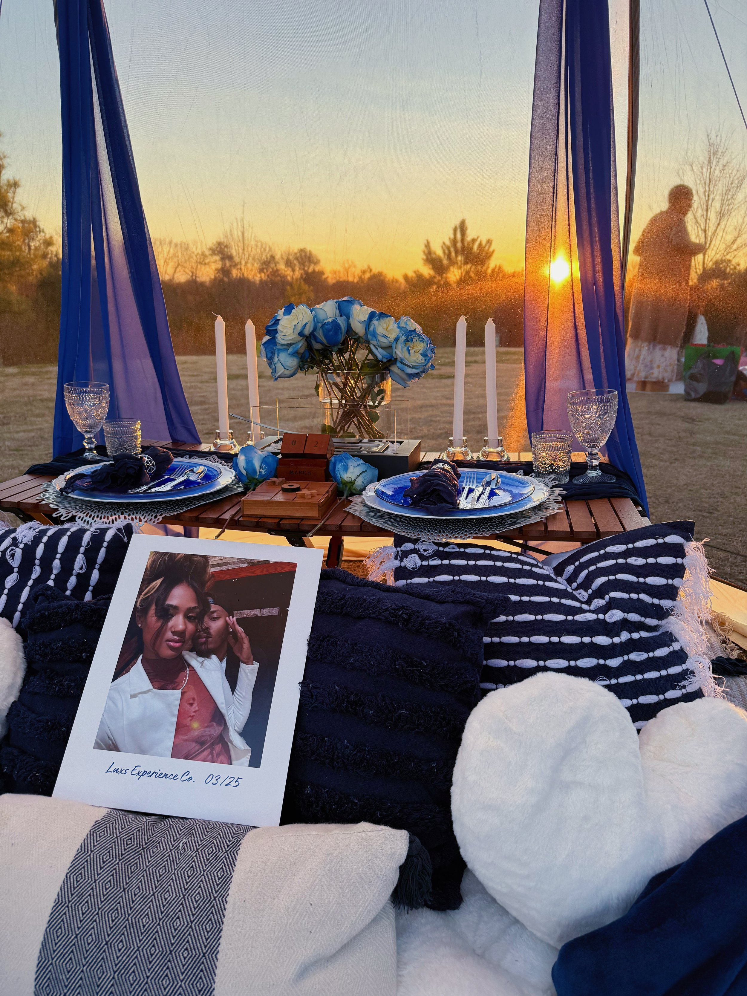 A romantic outdoor picnic setup at sunset with a table decorated with blue and white flowers, candles, and dinnerware, surrounded by pillows and blankets, and a photo of a woman in the foreground.