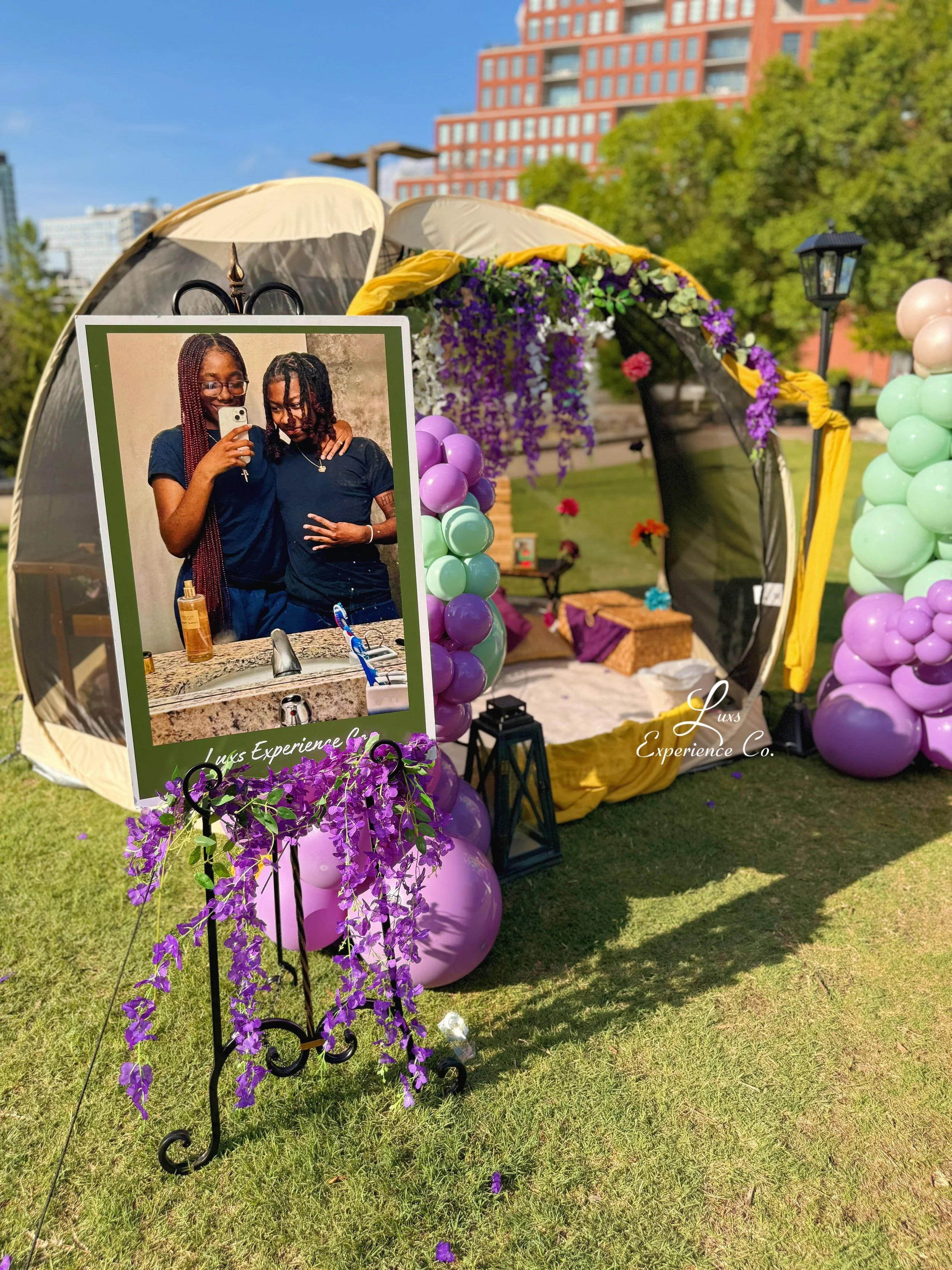 Decorative outdoor tent decorated with purple, green, and pink balloons and purple flowers, featuring a large reflective sign with two women taking a selfie in a bathroom mirror, surrounded by trees and buildings in the background.