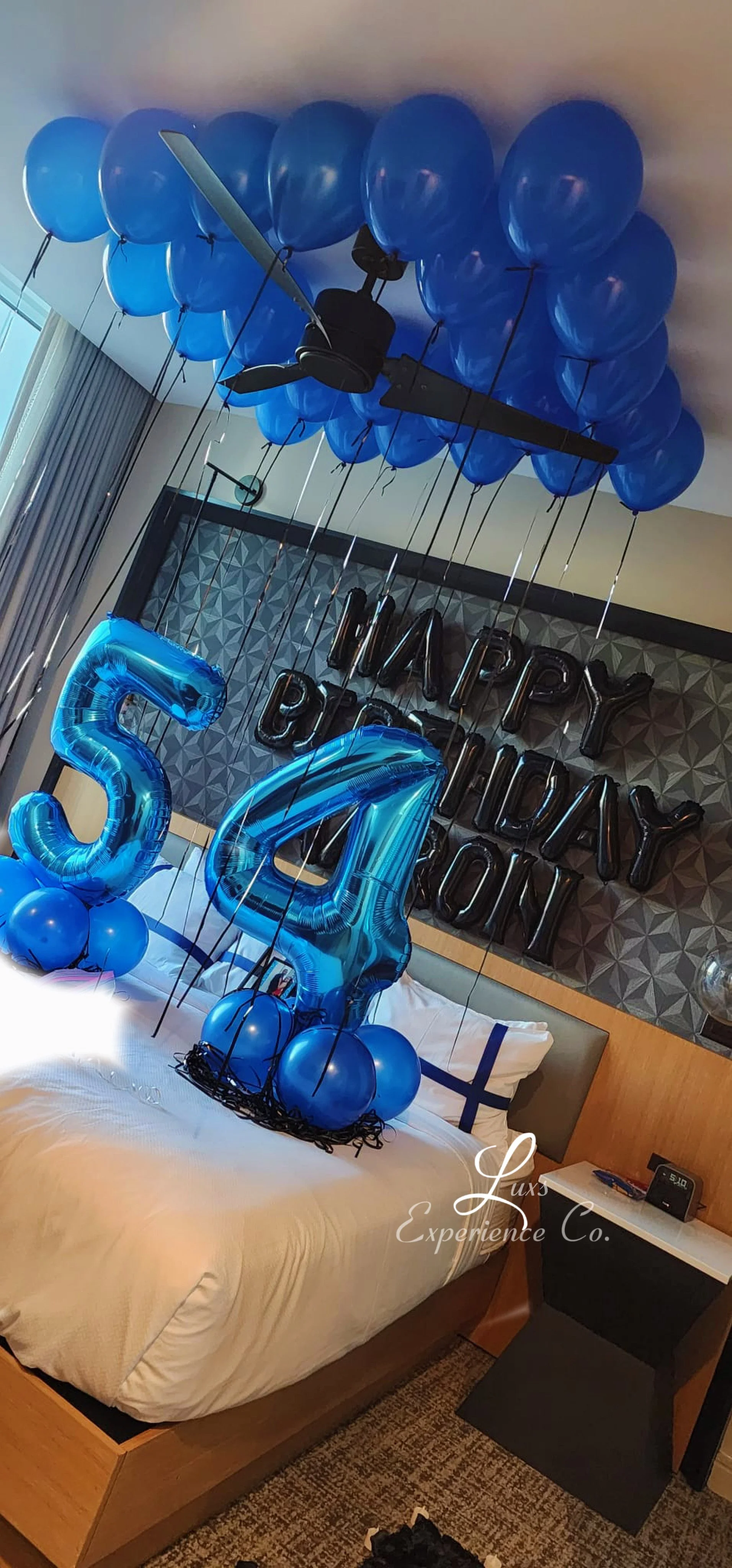 Hotel room decorated with blue balloons, including a large number 5 and 4, with a "Happy Birthday" sign on the wall, indicating a birthday celebration