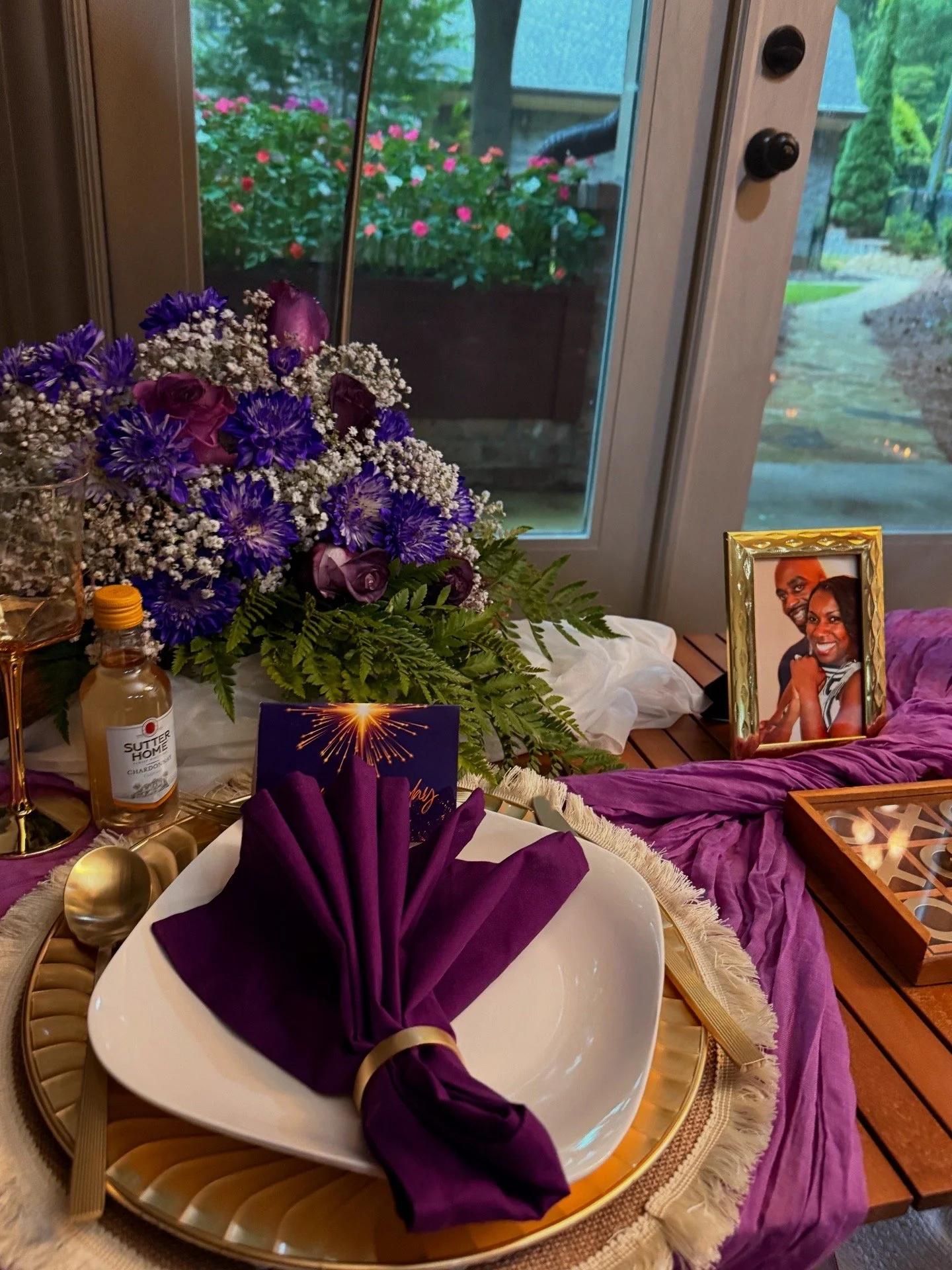Decorative table setup with a bouquet of purple and white flowers, a framed photo of a couple, a purple napkin folded like a fan, and a wedding card. The scene is near a glass door with a garden outside.