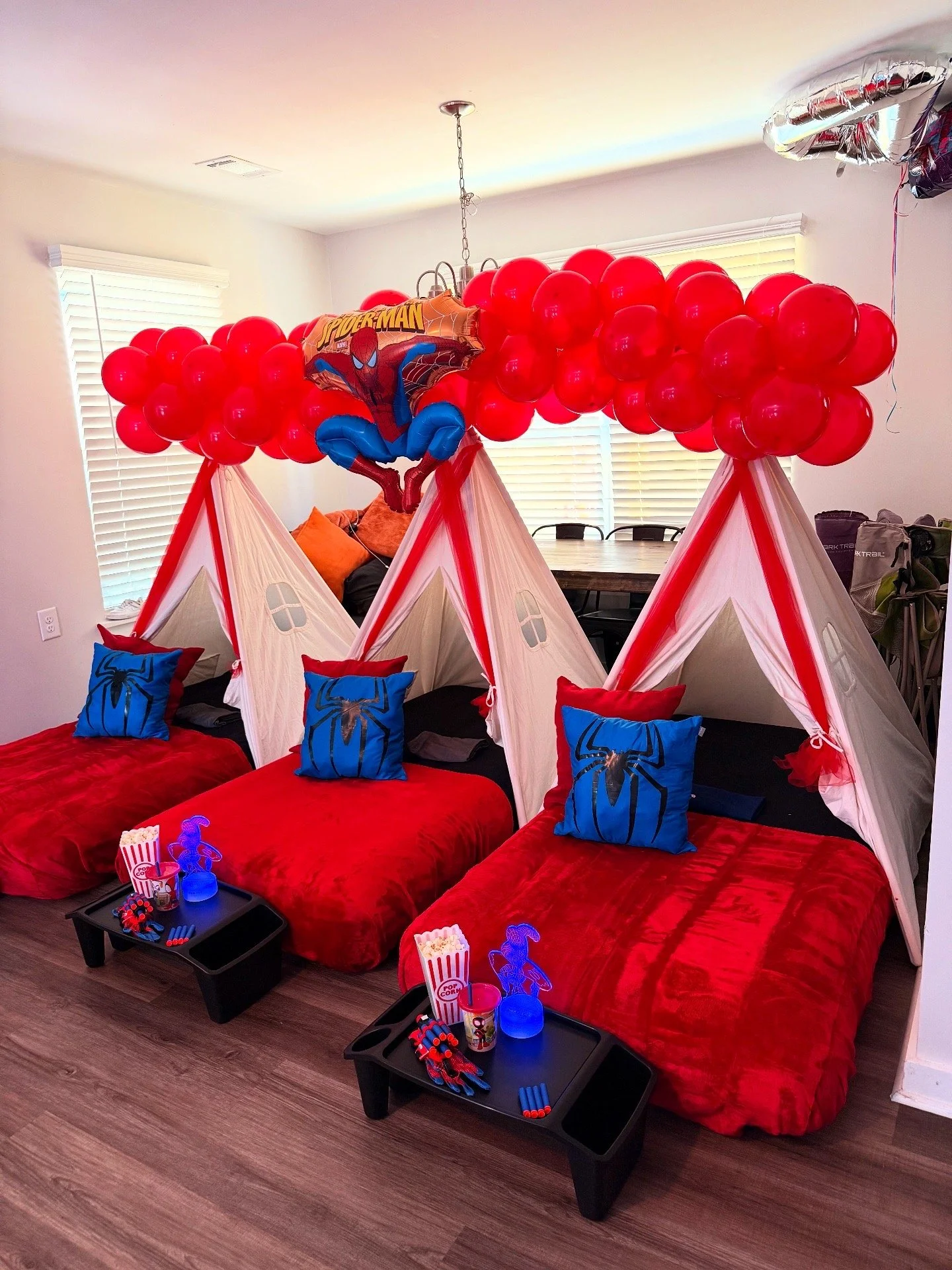 Three beds with Spider-Man themed pillows and red blankets, decorated as tents with white fabric and red accents, topped with red balloons and a Spiderman balloon, set up for a children's party.