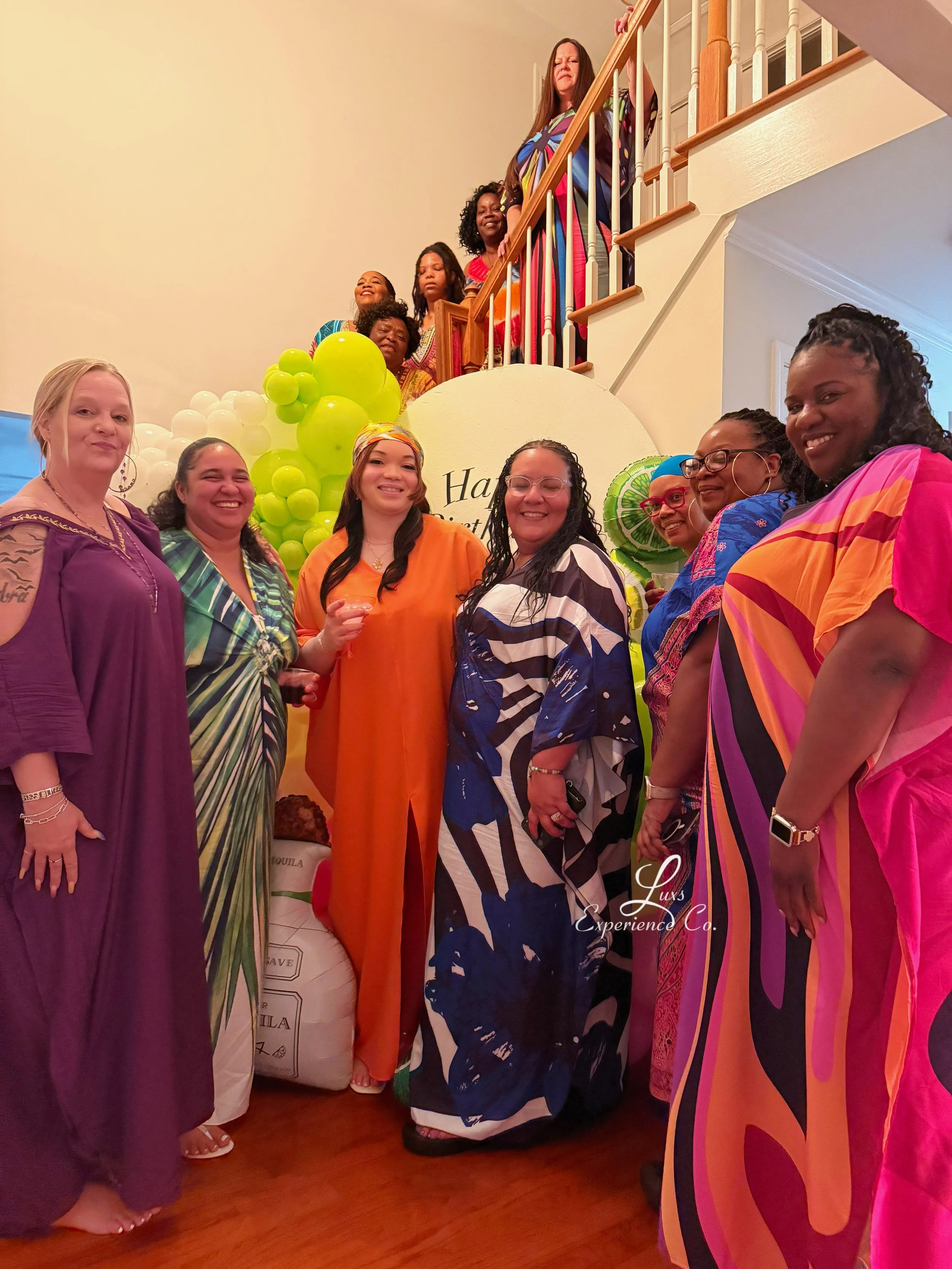 A group of women at a celebration, gathered indoors. They are dressed in colorful, patterned clothing, and are standing in front of a balloon decoration and a large sign that says 'Happy'. Some women are smiling and holding drinks. The setting appear