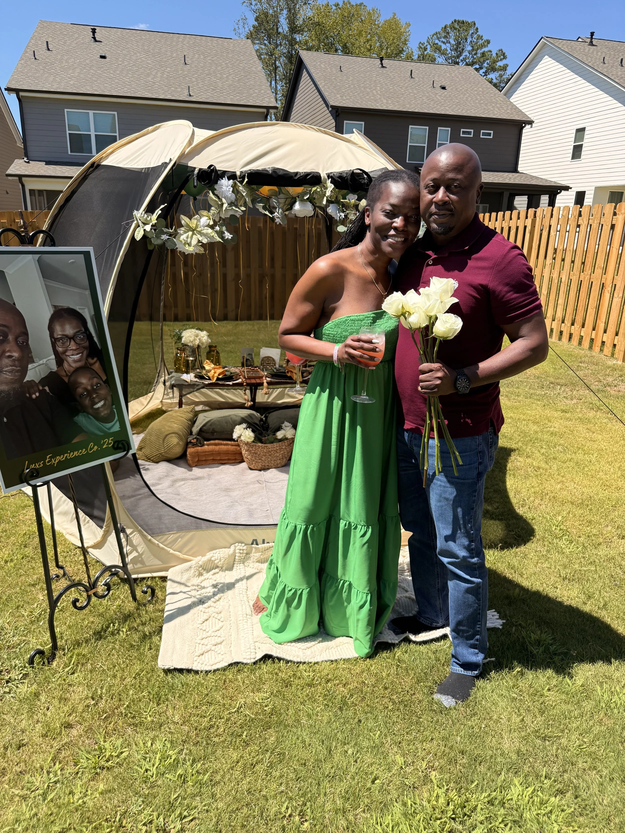 A happy couple standing in their backyard around a decorated small tent, holding flowers and a beverage, with a photo board showing pictures of them, under a clear blue sky.