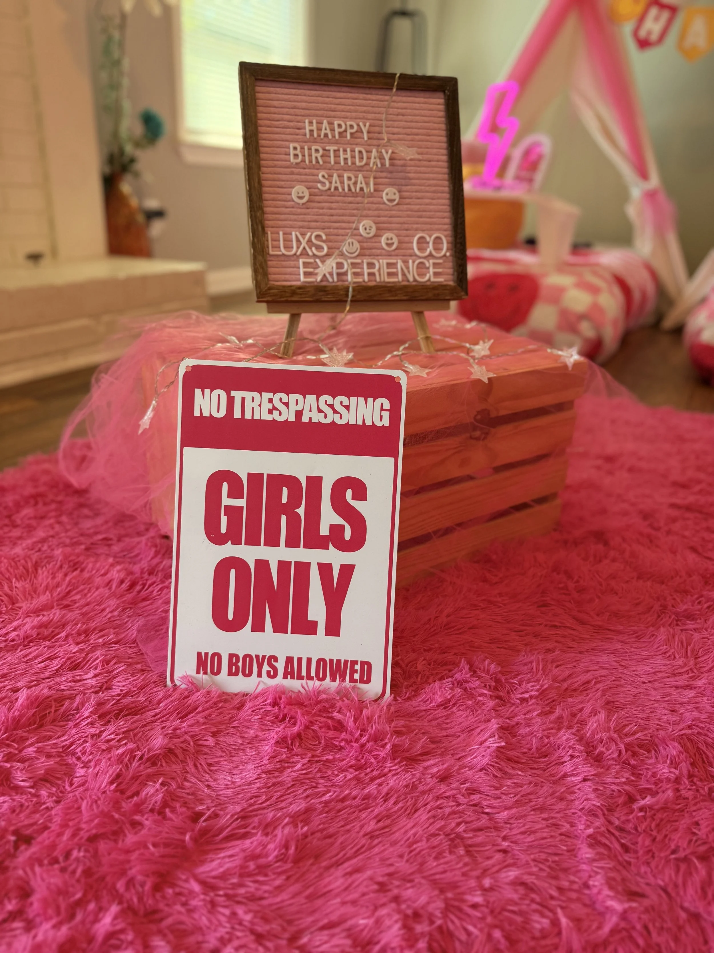 A pink birthday setup with a sign that says 'Girls Only, No Boys Allowed' placed on a pink fluffy rug, with a decorated bed in the background featuring pink and red pillows and a pink tent with lightning bolt decoration.