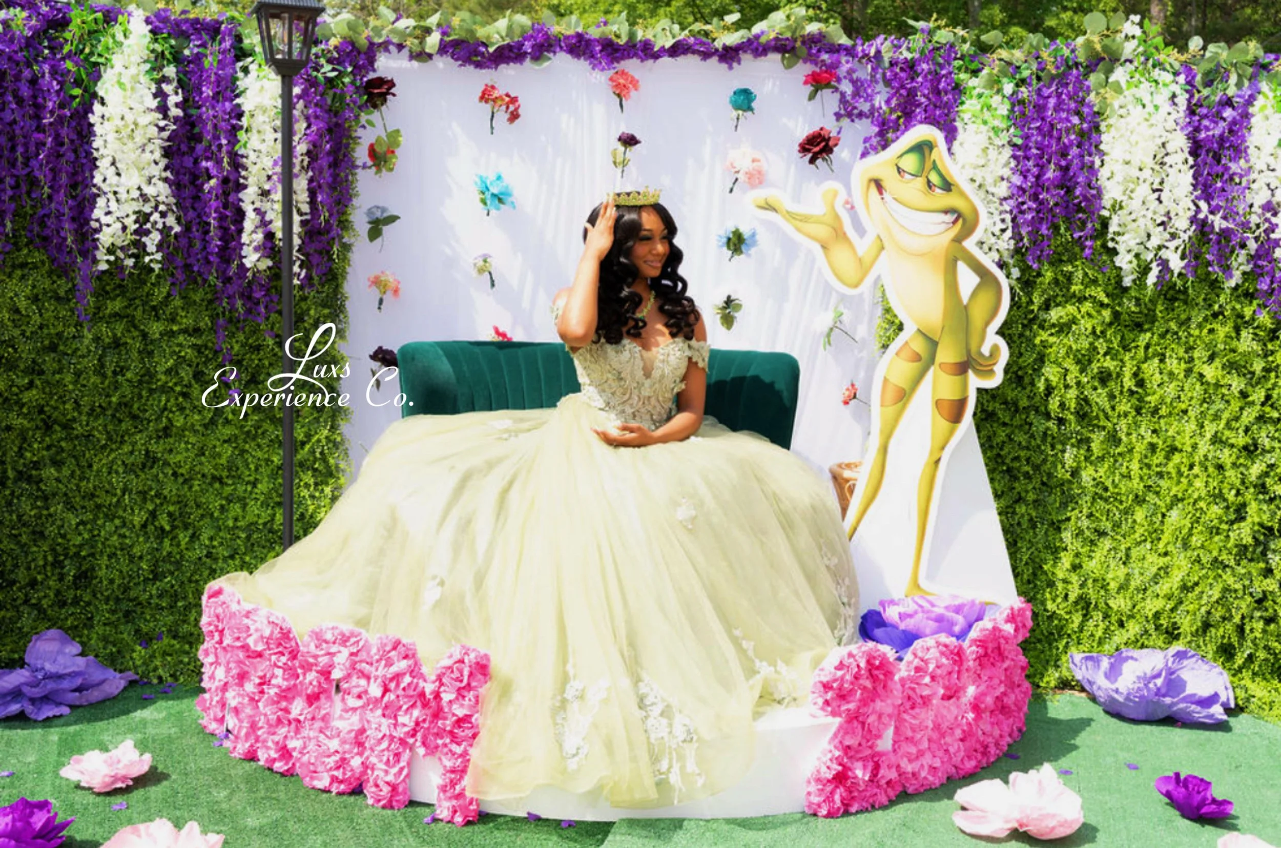 A woman dressed in a light yellow gown sitting on a green velvet couch, wearing a tiara, surrounded by colorful flowers, with a backdrop of purple and white flowers and a cartoon character of a smiling yellow creature with stripes, pointing and gestu