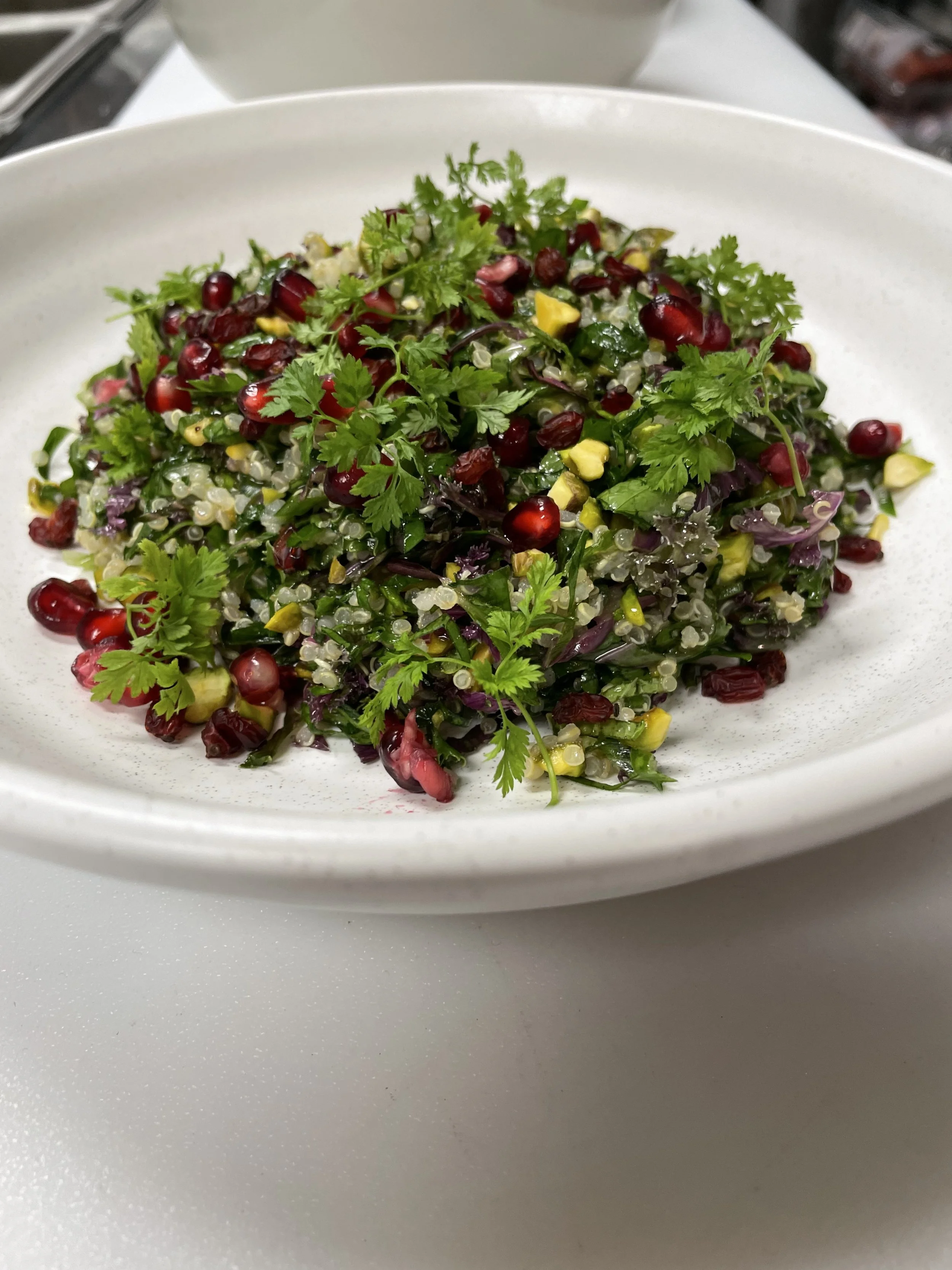 A white bowl filled with a colorful quinoa salad, garnished with pomegranate seeds, chopped avocado, herbs, and leafy greens.