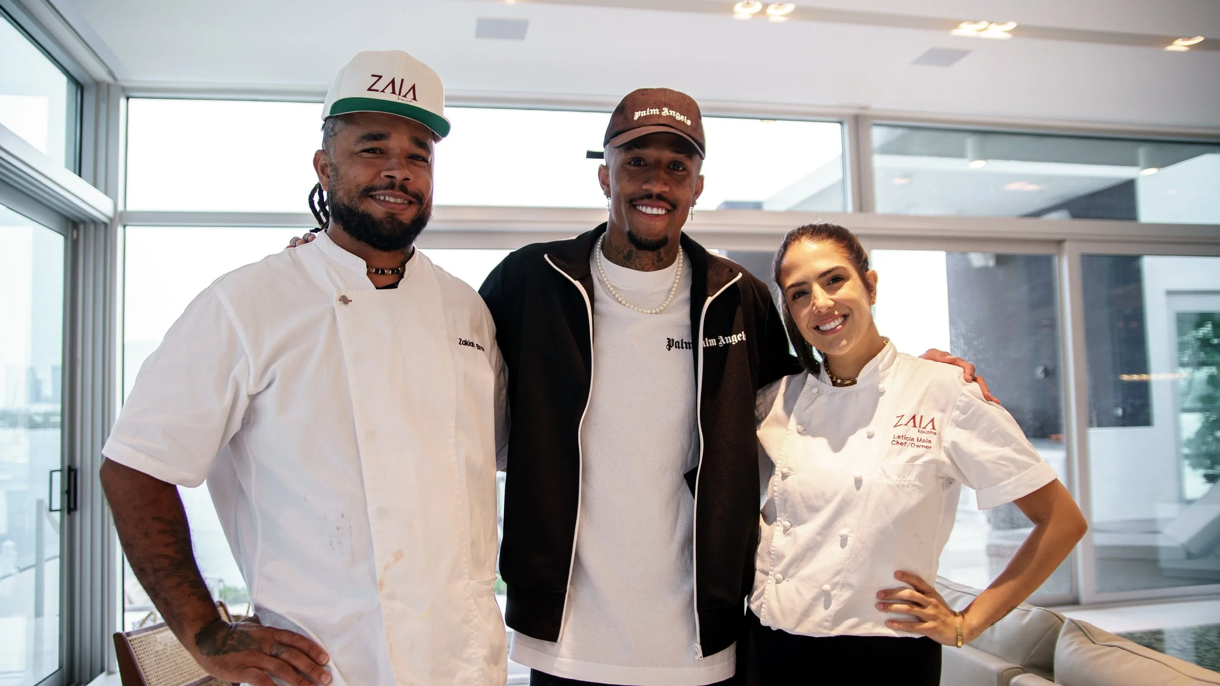 Three people standing together indoors, smiling, with large windows in the background. The person on the left is wearing a white chef's coat and a white cap. The person in the middle is dressed in casual clothing with a jacket and a baseball cap. The person on the right is wearing a white chef's coat with 