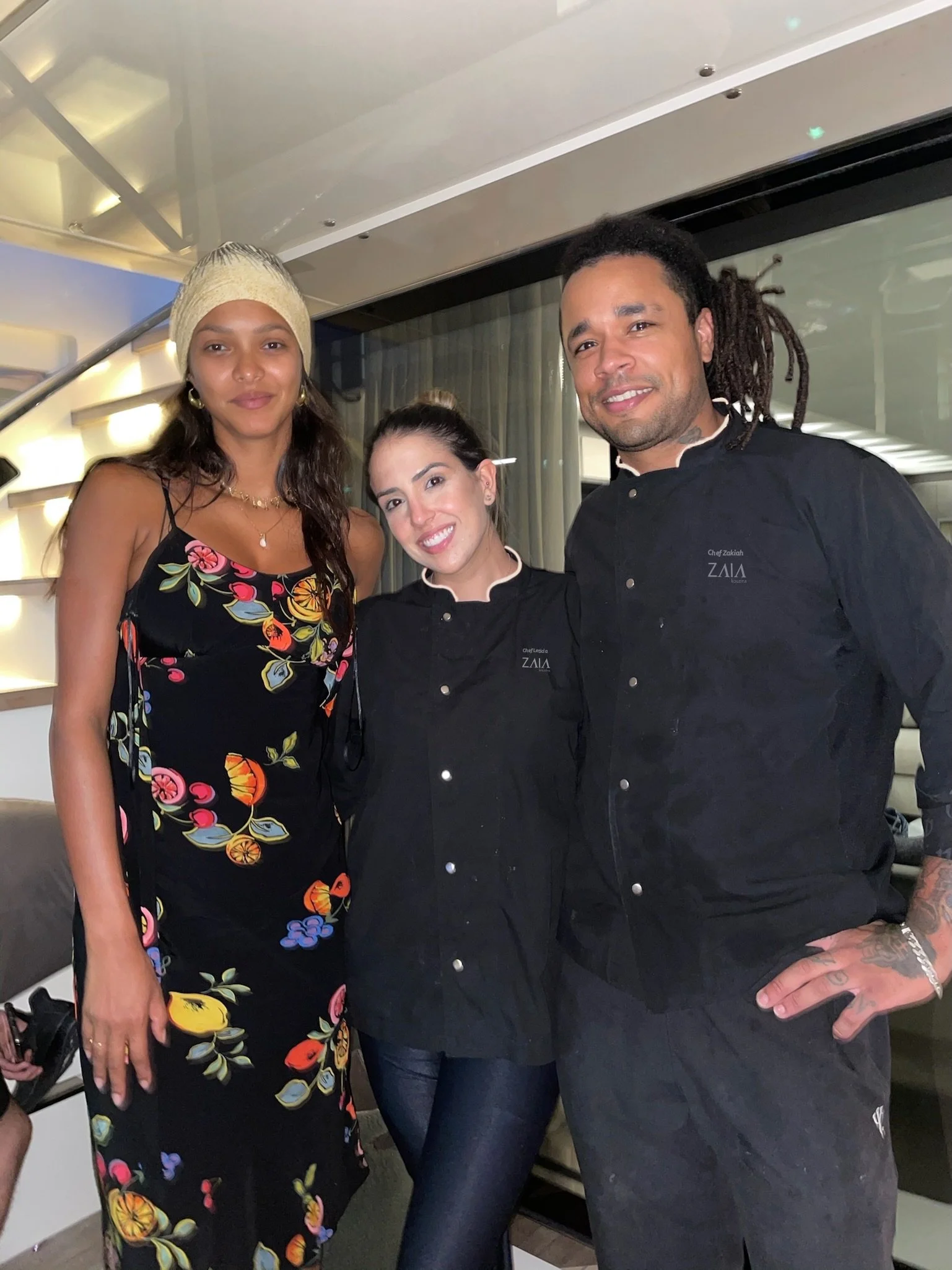 Three people, two women and one man, posing together indoors. The woman on the left is wearing a floral dress and a beige head covering. The woman in the middle is wearing a black chef's coat, smiling. The man on the right has dreadlocks and is dressed in a black chef's coat, also smiling.