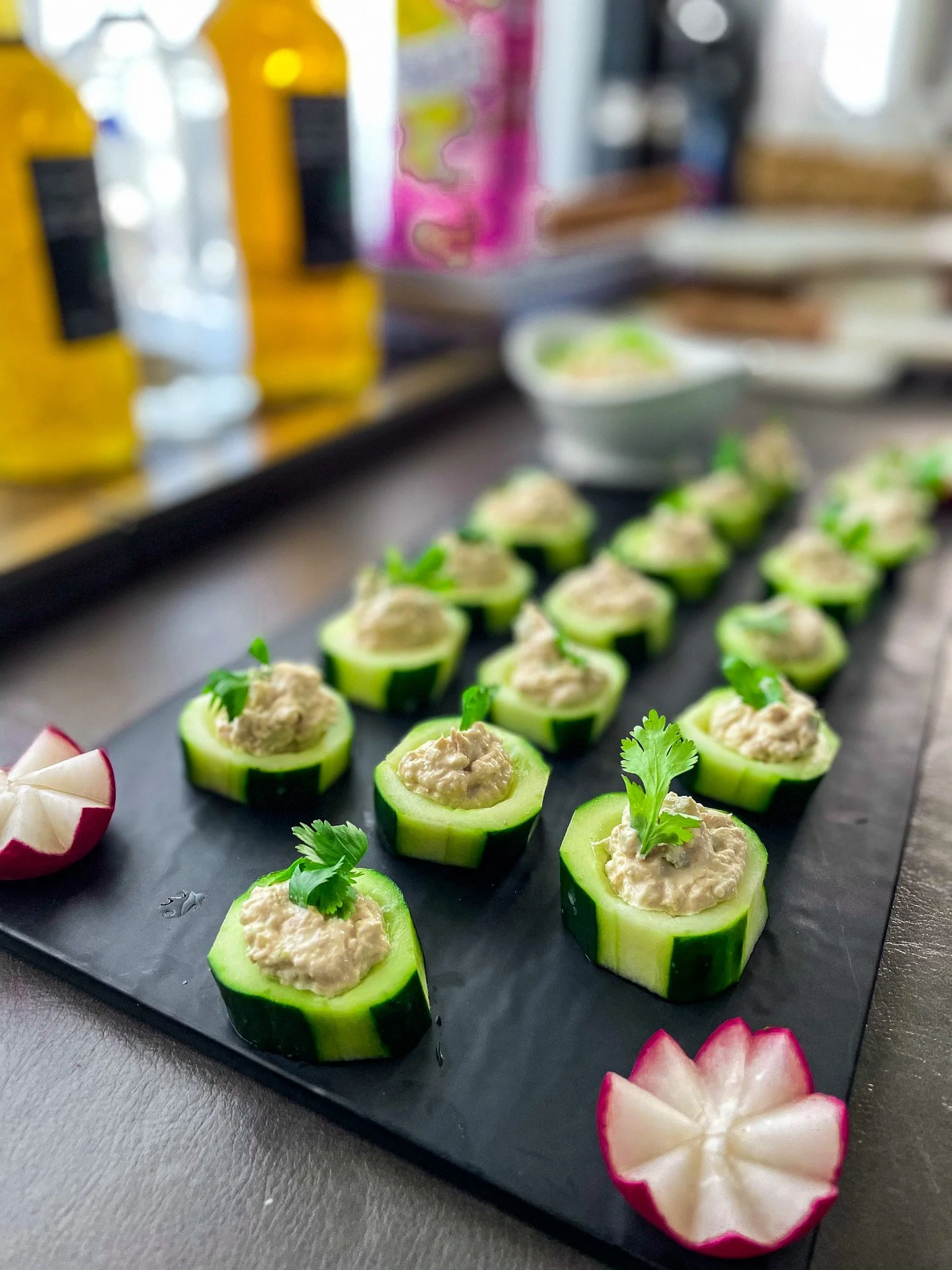 Cucumber canapés topped with a creamy spread and garnished with cilantro on a black serving tray, with radish flowers at the corners, blurred bottles and bowls in the background.