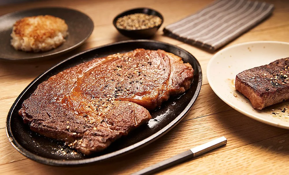 Raw and cooked steaks on plates with seasoning, a small bowl of black pepper, and a side dish.