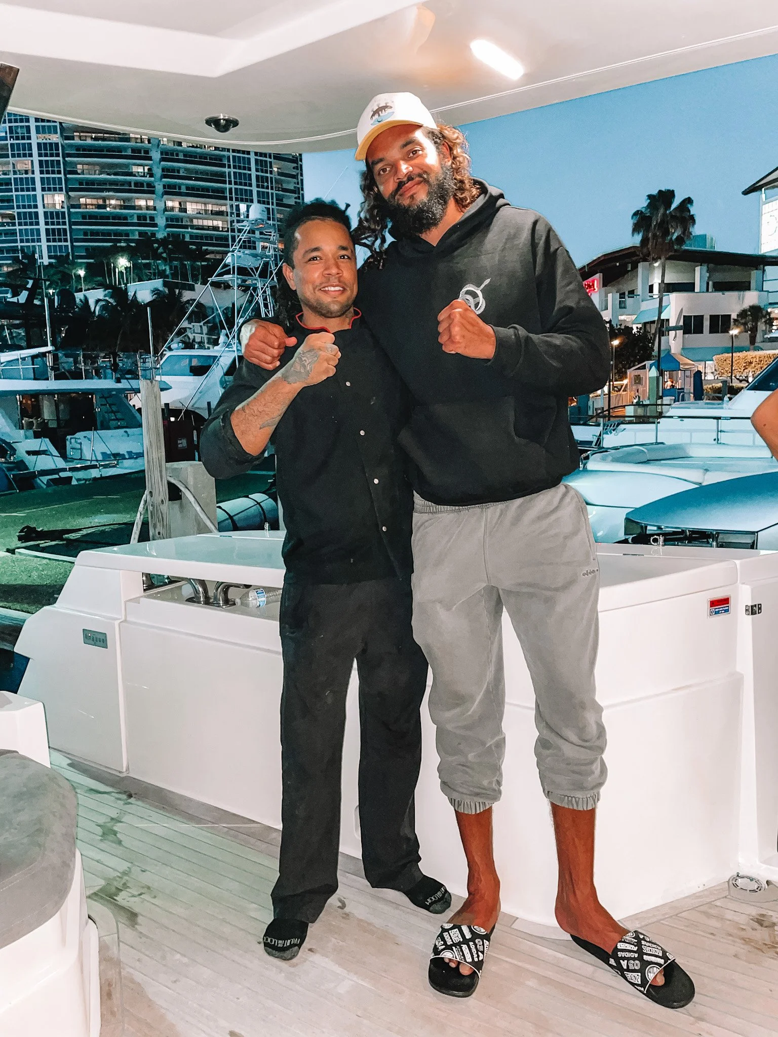 Two men standing side by side on a boat dock at a marina during evening. They are wearing casual attire, with one in a black button-up shirt and the other in a black hoodie, both with fists raised. The taller man is barefoot, wearing grey sweatpants and slides, while the shorter man has sneakers. Background shows boats, modern buildings, and palm trees.