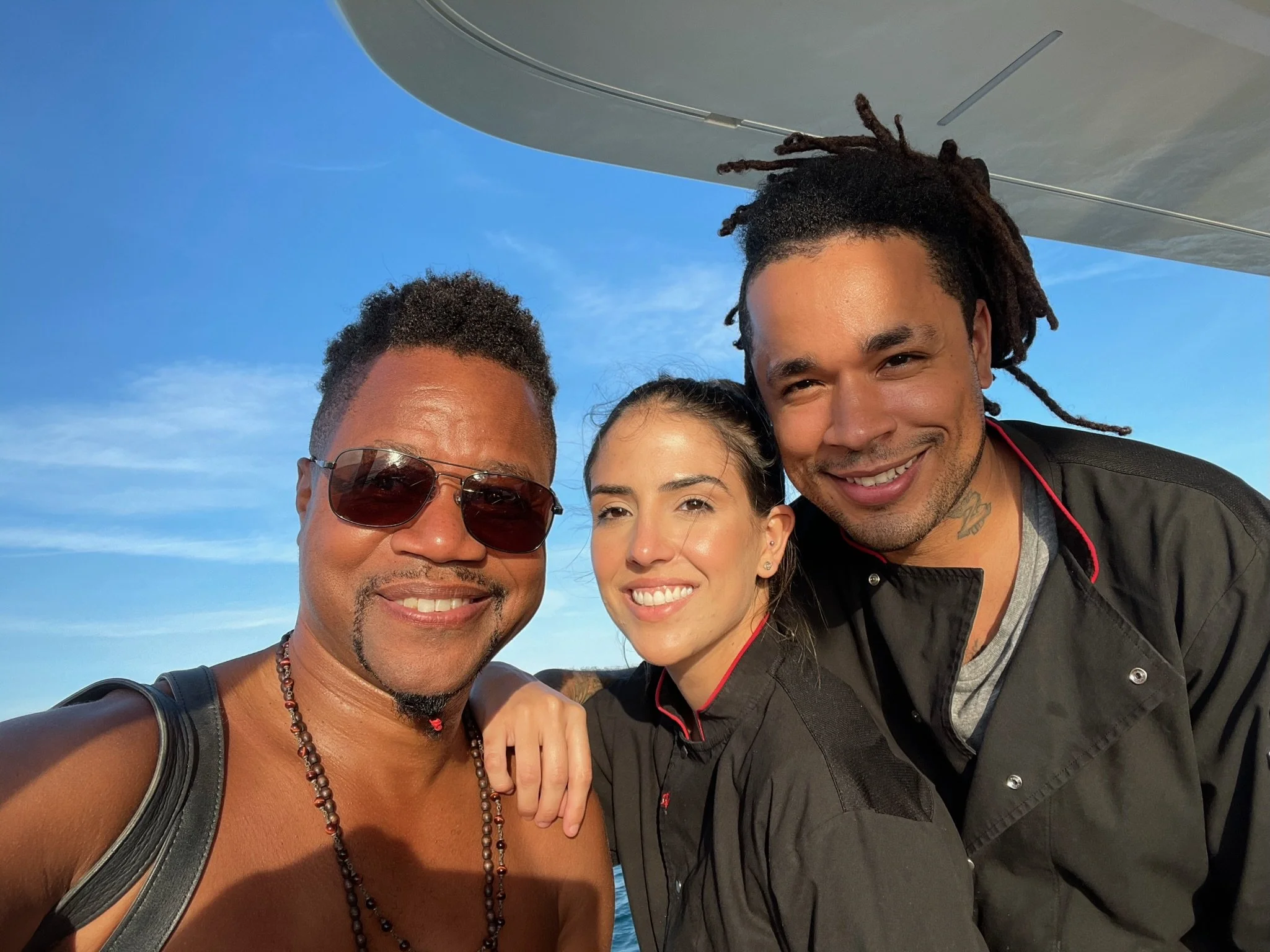 Three friends smiling for a selfie outdoors, with a blue sky, ocean, and part of a boat roof visible.
