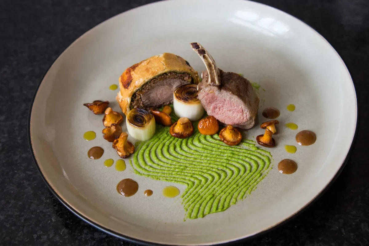 A plate of gourmet food featuring a cooked lamb chop, a slice of veggie stuffing, miniature grilled onions, mushrooms, green pea puree with a finger-shaped piped pattern, and scattered sauce dots.