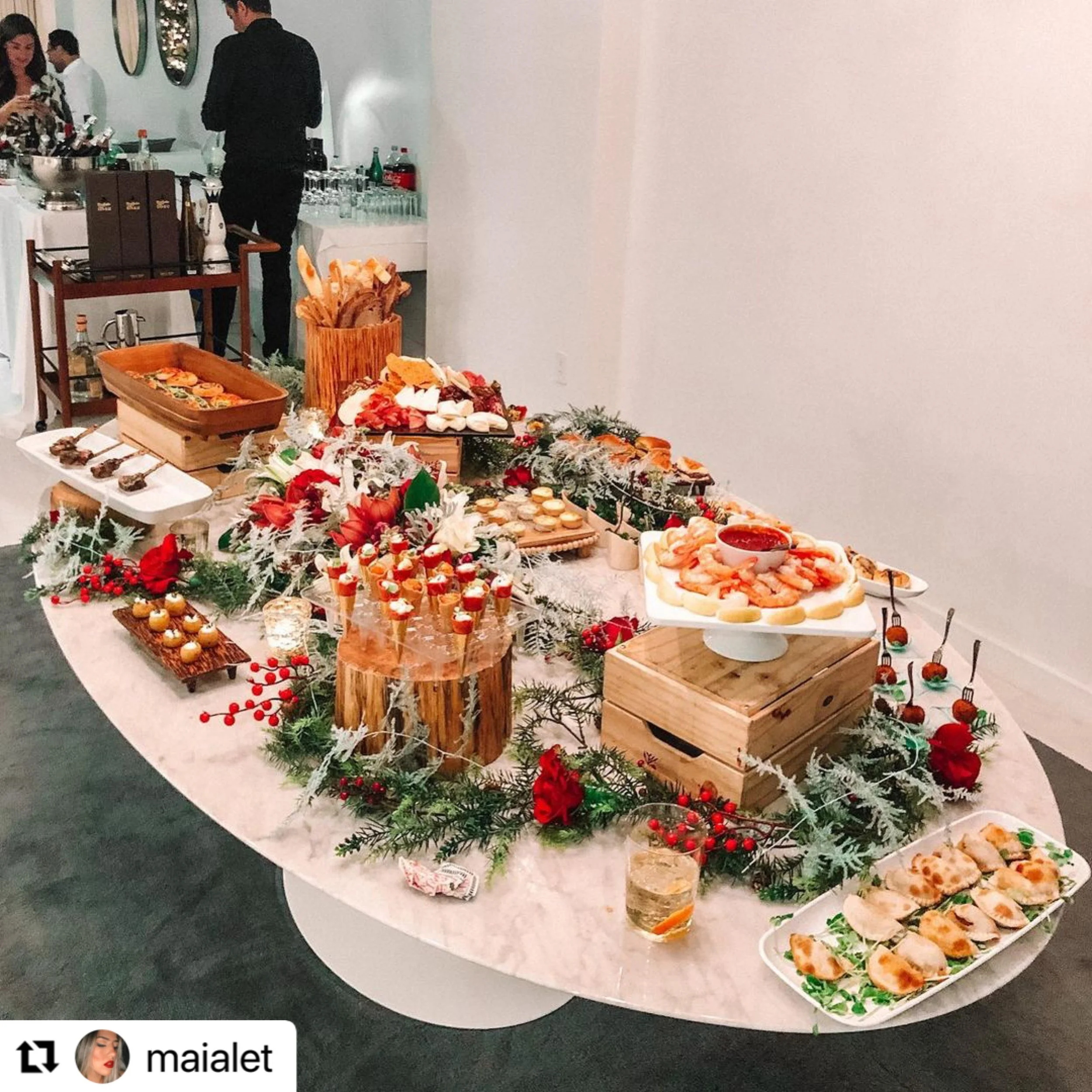 Spread of holiday-themed appetizers and desserts on a decorated table with red and green foliage, berries, and red flowers. The table includes cheese, shrimp, bite-sized snacks, and sweets, with some items on wooden and ceramic dishes.