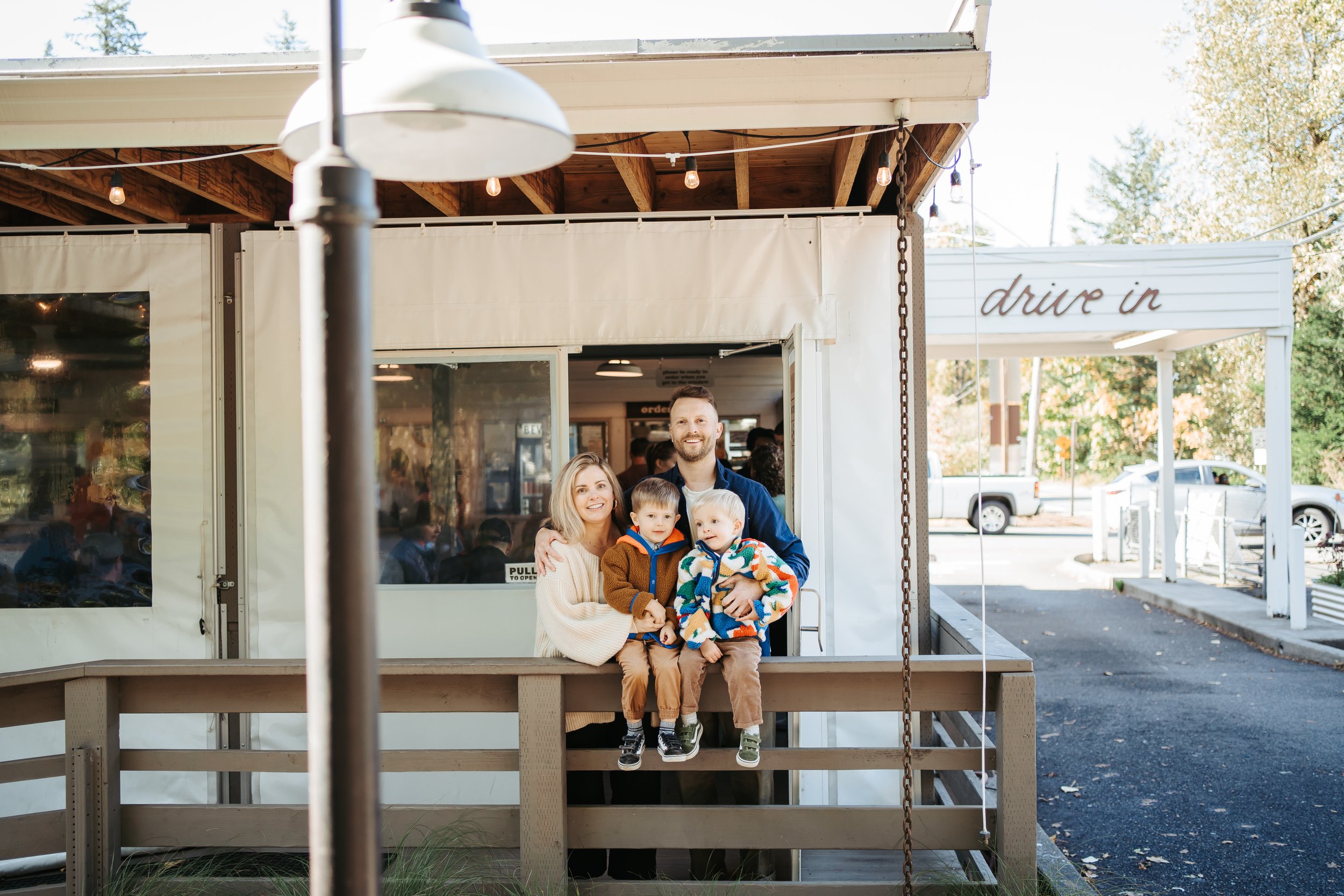 A family of four standing on the porch of a restaurant with a drive-in sign in the background, smiling at the camera.