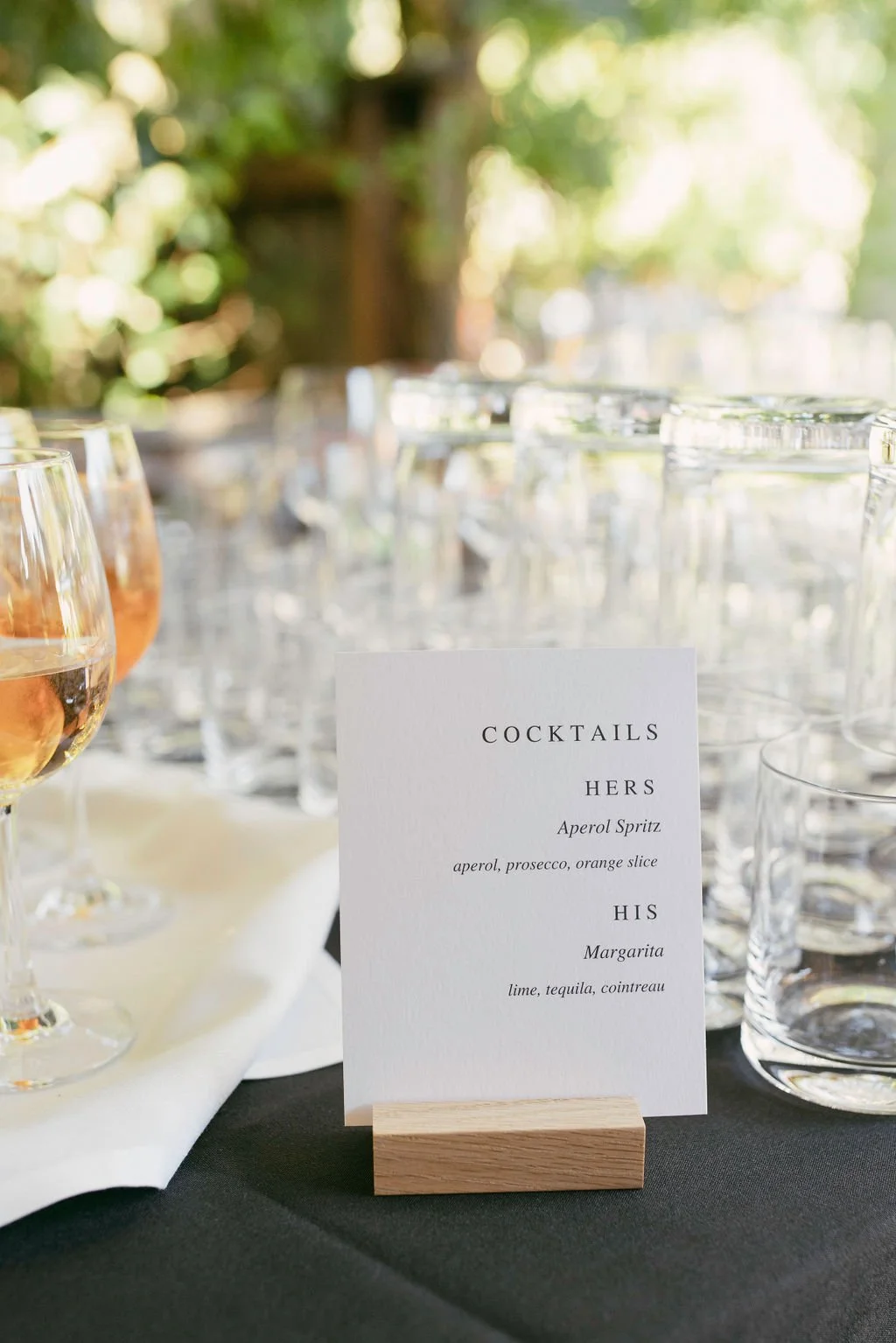 Table setting with glasses of rosé wine, a menu card titled 'Cocktails', listing 'Hers' with Aperol Spritz and 'His' with Margarita, on a black and white tablecloth with a blurred outdoor garden background.