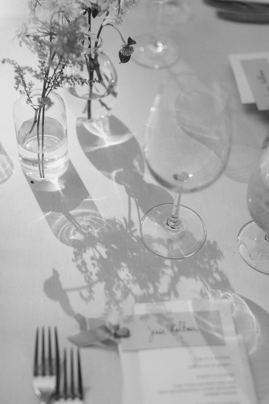 A table setting with a flower arrangement in a glass vase, a wine glass, a fork, and a place card on a table covered with a white tablecloth. The shadows of the flowers and vase are visible on the table.