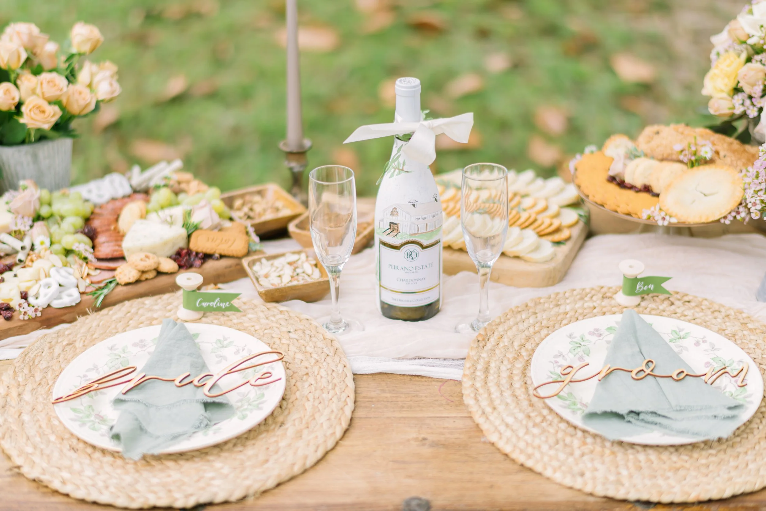 Outdoor table set for a celebration with a bottle of wine, two flutes, cheese, crackers, cookies, and flowers, with a place card reading 'Grandma'.