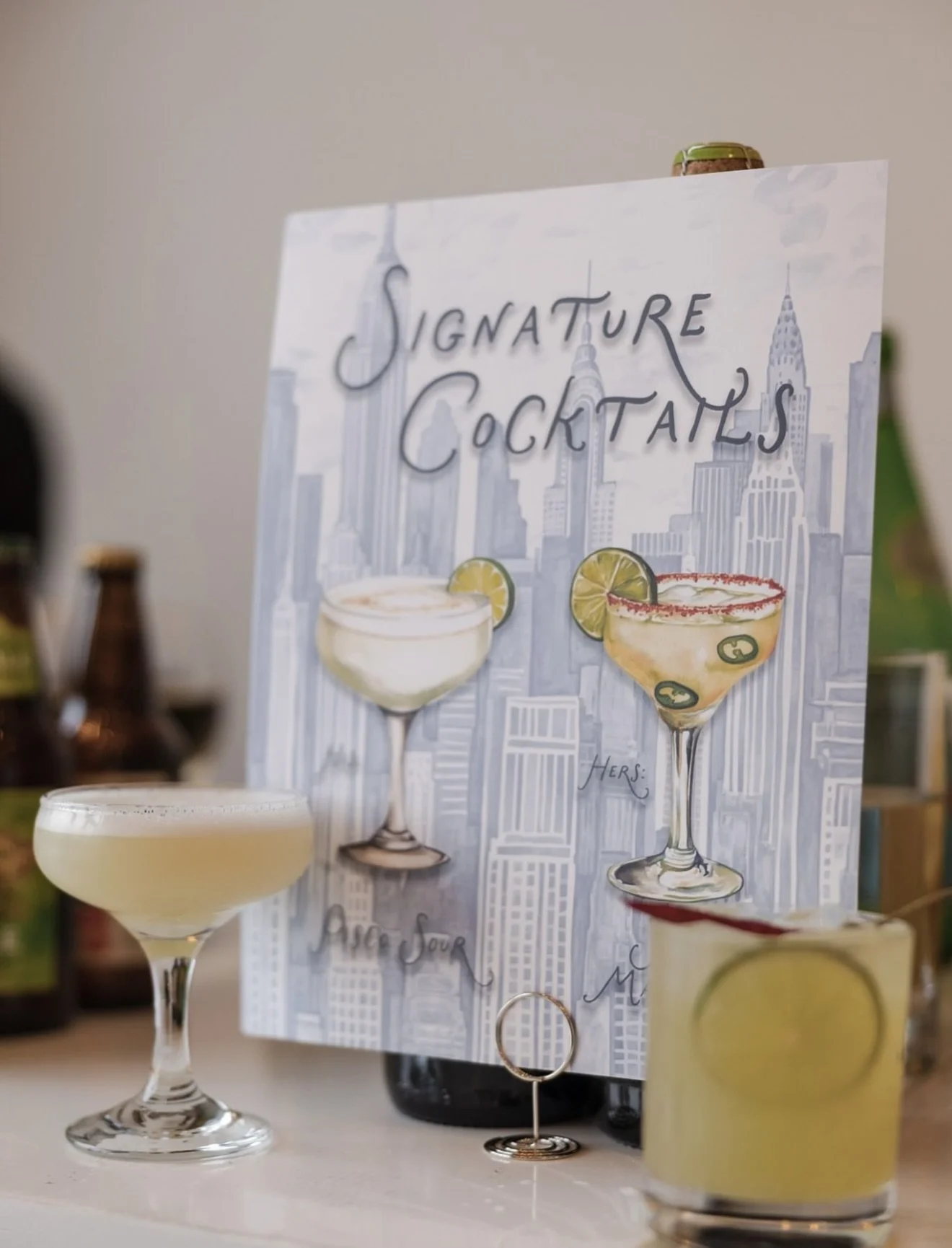 Display of signature cocktails on a table with a sign featuring illustrations of cocktails and a New York City skyline in the background.
