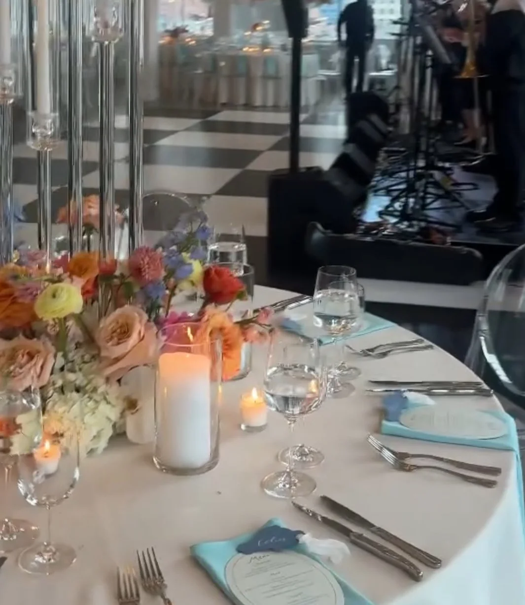 Elegant banquet table with floral centerpiece and candles, set with glassware, cutlery, and menus, at a formal event or wedding reception with a stage and band in the background.